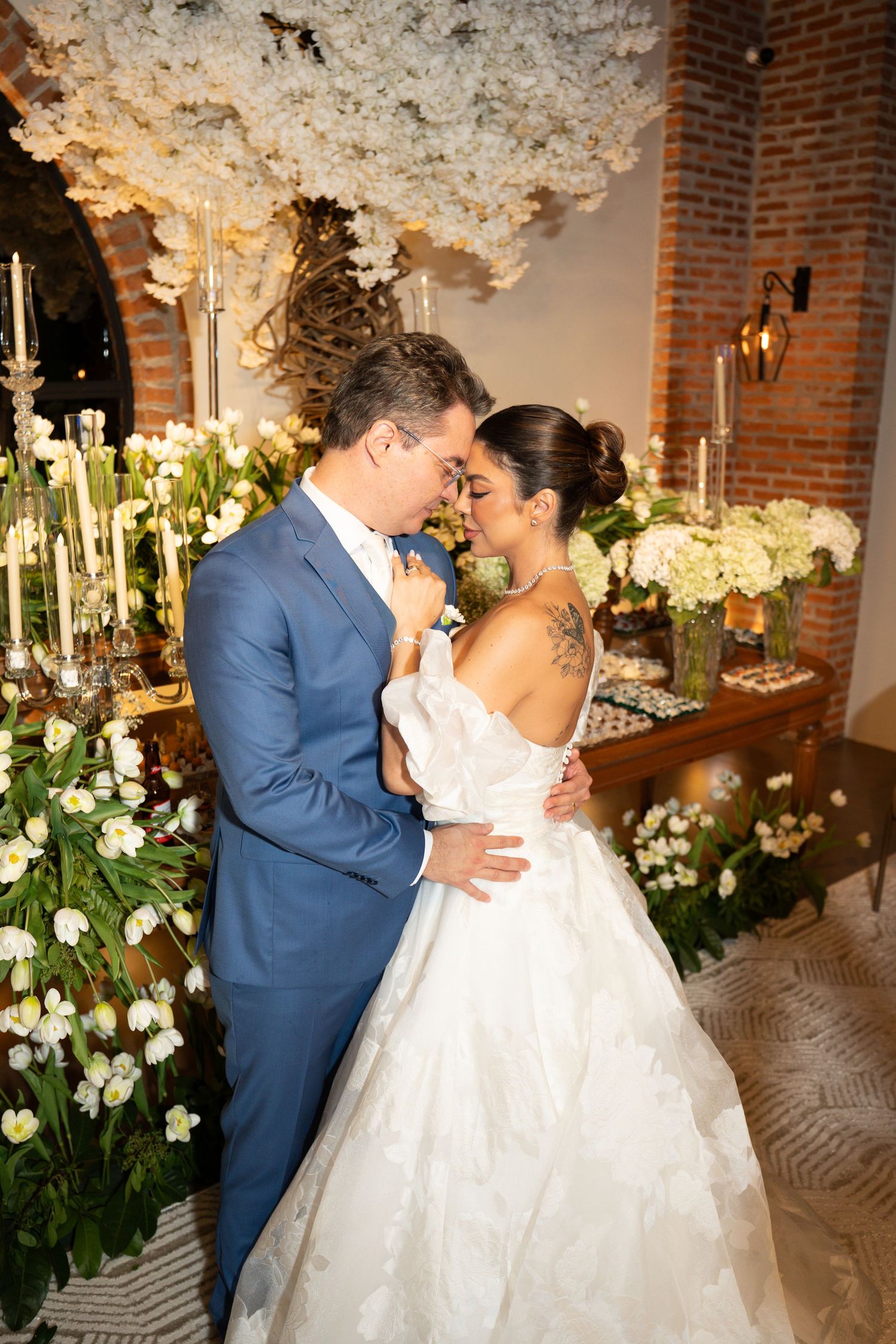 Bride and groom share romantic moment in wedding attire, surrounded by lush white floral arrangements and candlelight.