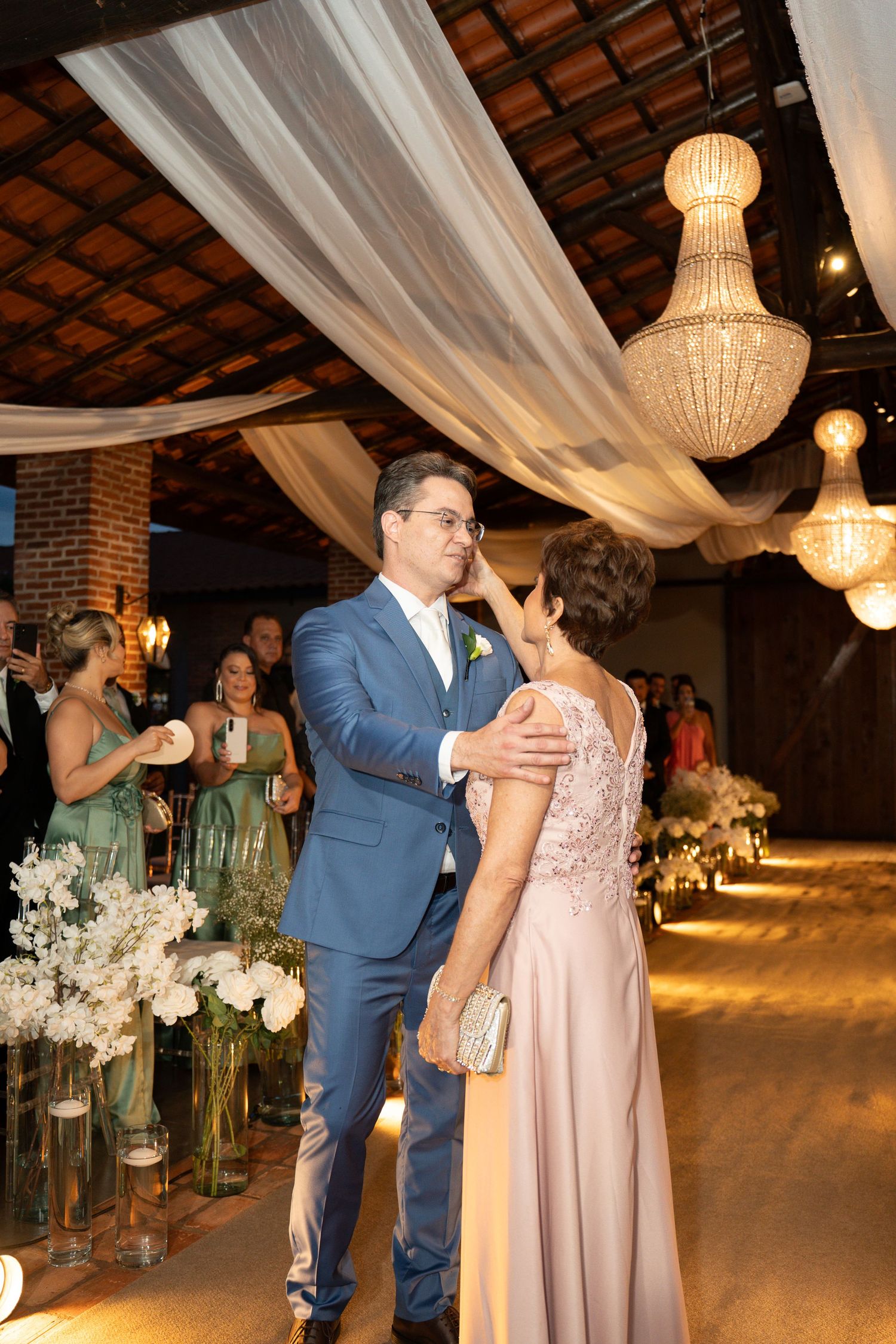 Groom in blue suit shares a tender moment with a woman in pink lace gown at elegant wedding reception with chandeliers.