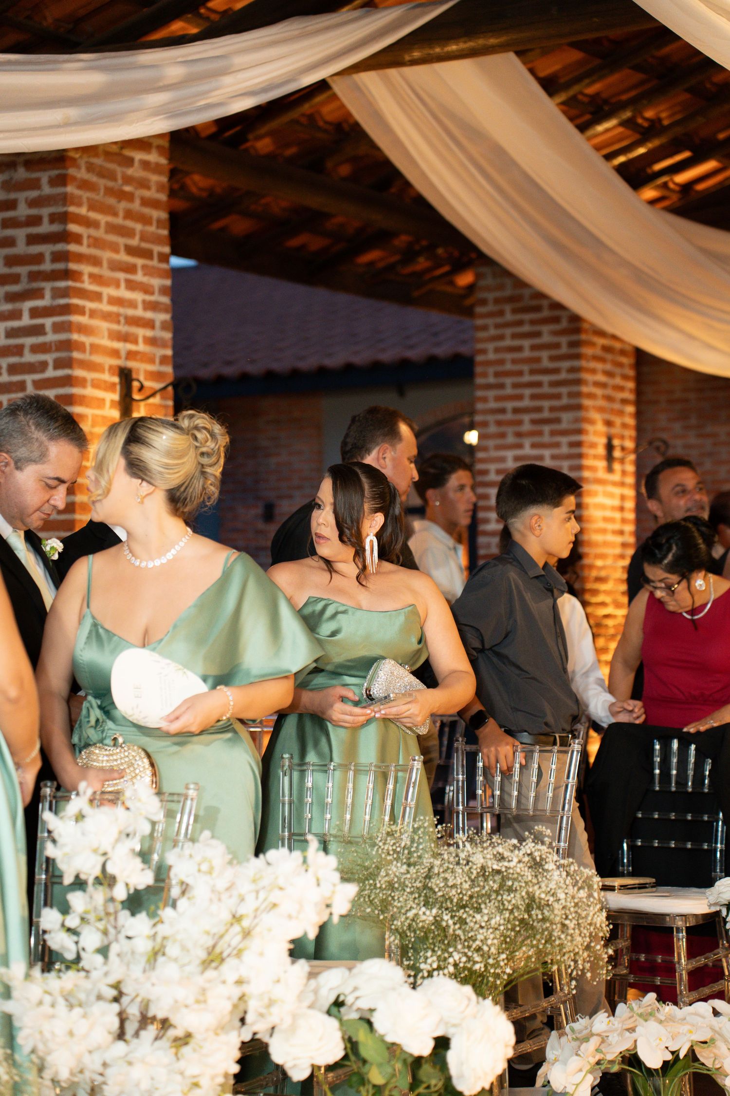 Bridesmaids in sage green dresses stand at a wedding reception in a rustic brick venue with white floral arrangements.
