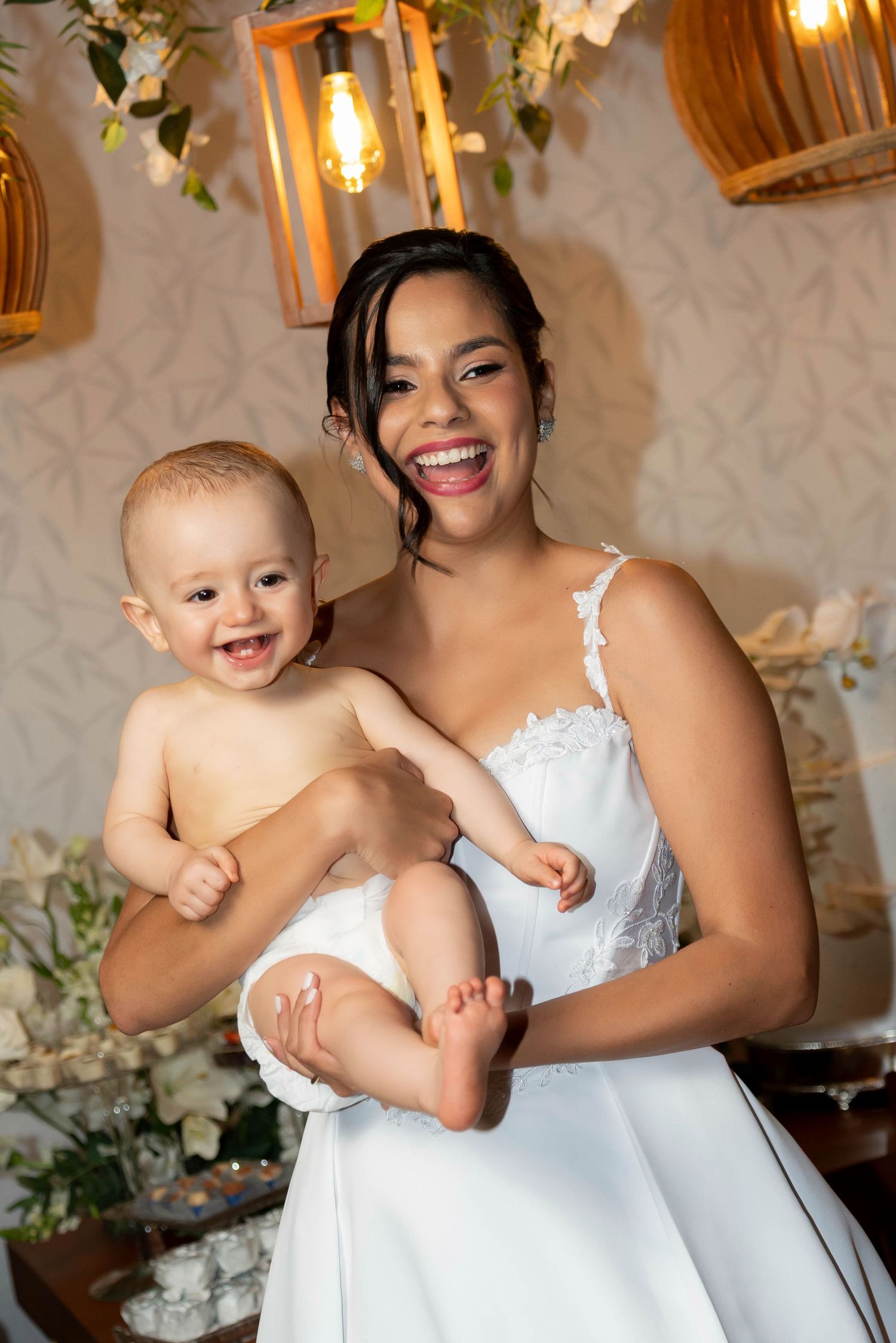 A joyful bride in a white dress holds a smiling baby at a decorated celebration venue.