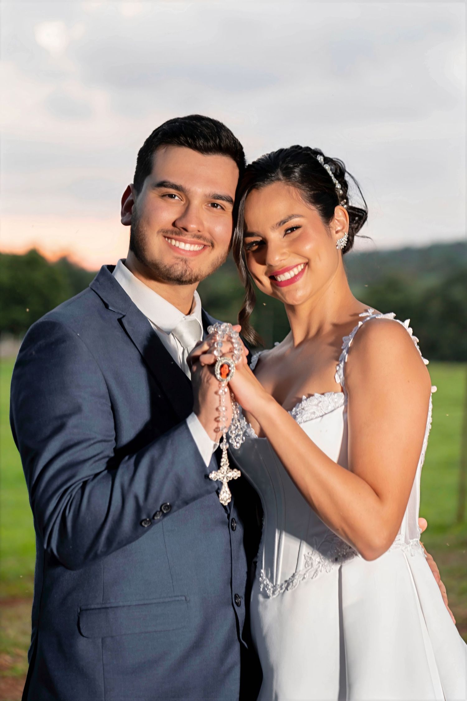 Smiling bride and groom holding a rosary together outdoors at sunset, wearing wedding attire.