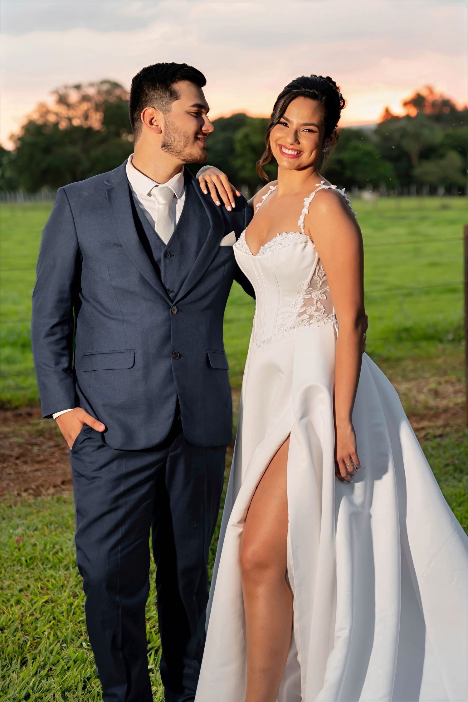 A couple in wedding attire poses outdoors at sunset, groom in navy suit, bride in white lace gown with slit.
