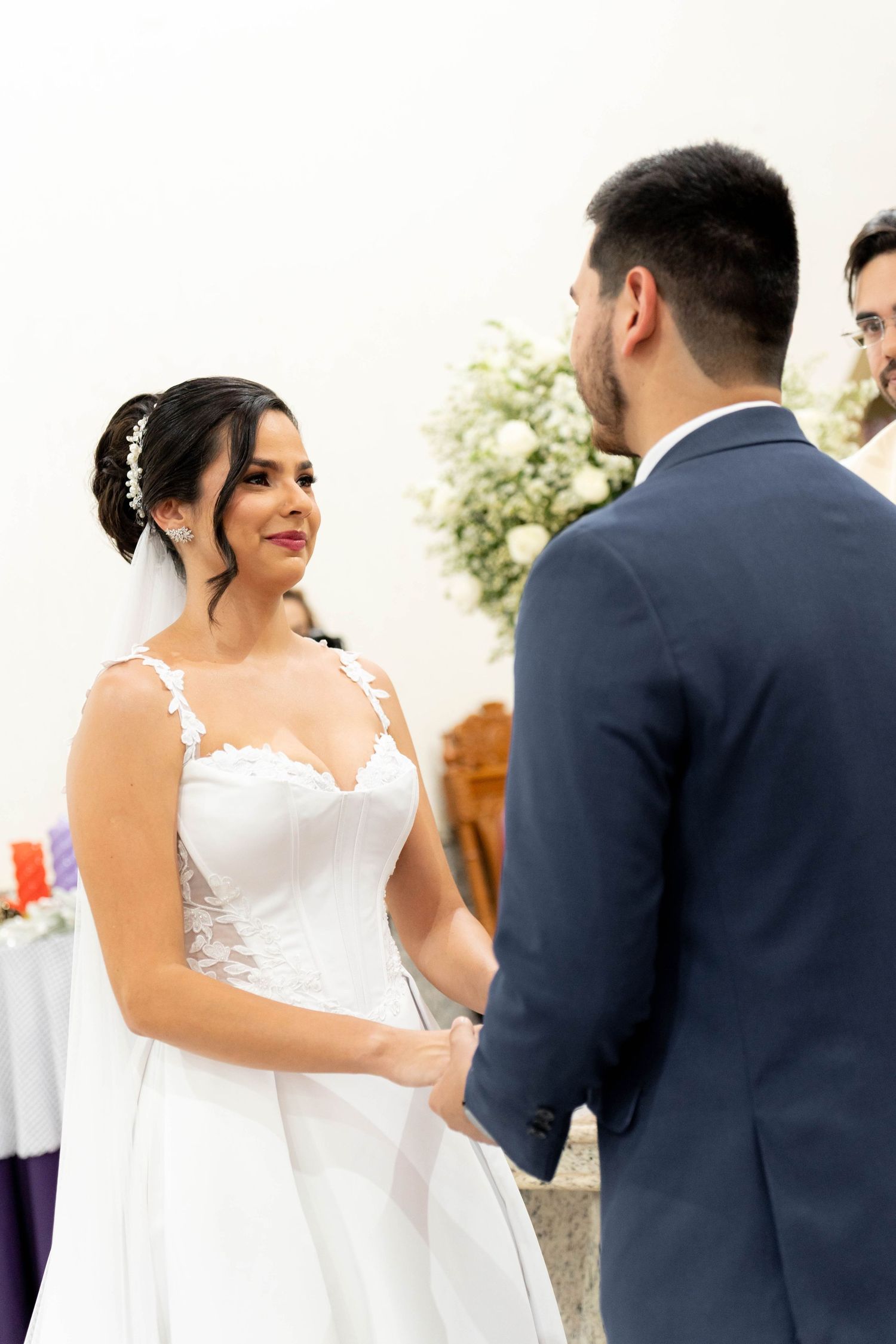 Bride in white lace gown smiling at groom in navy suit during wedding ceremony, holding hands at the altar.