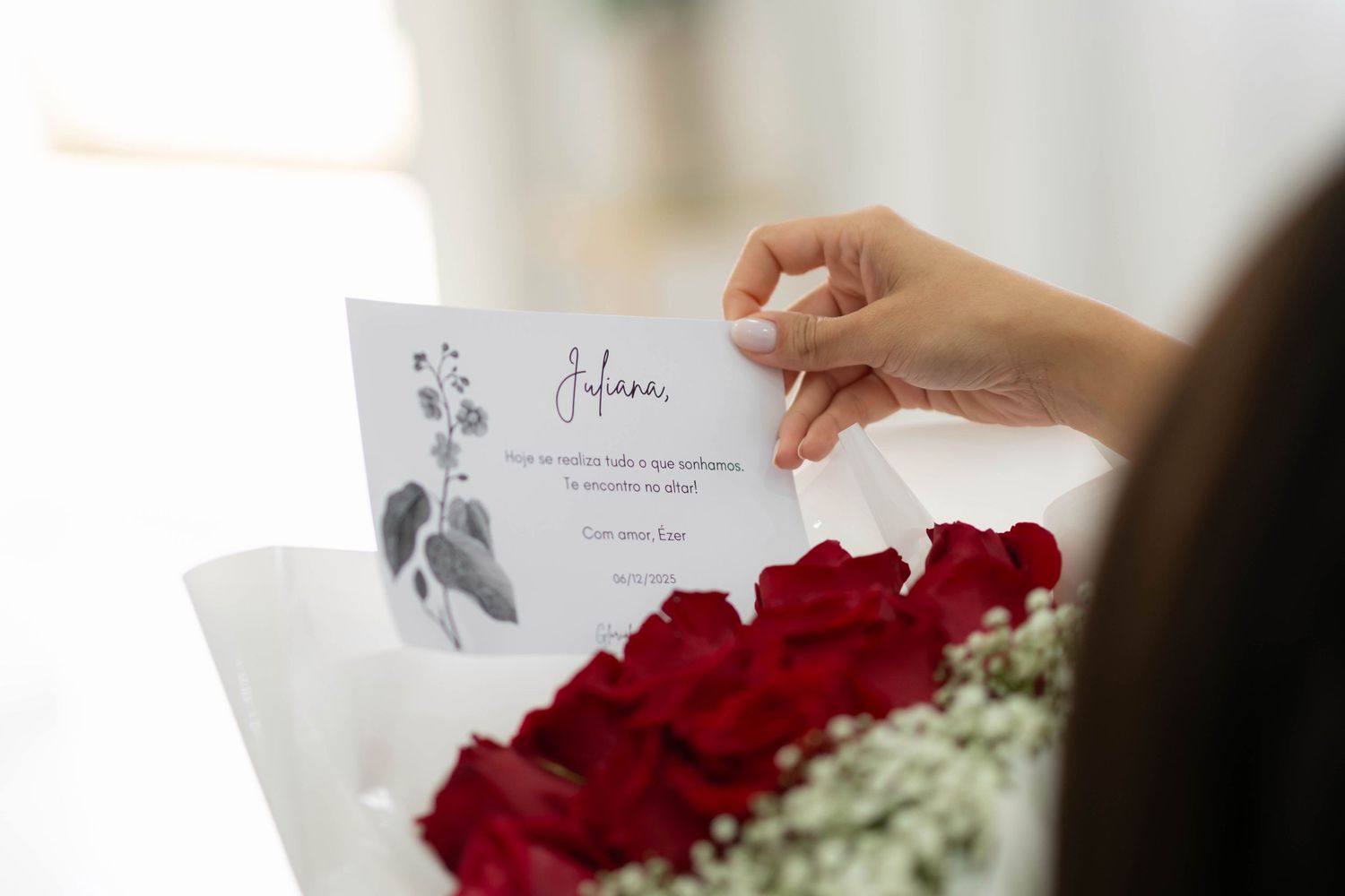 A hand holds a personalized card above a bouquet of red roses wrapped in white paper with baby's breath.