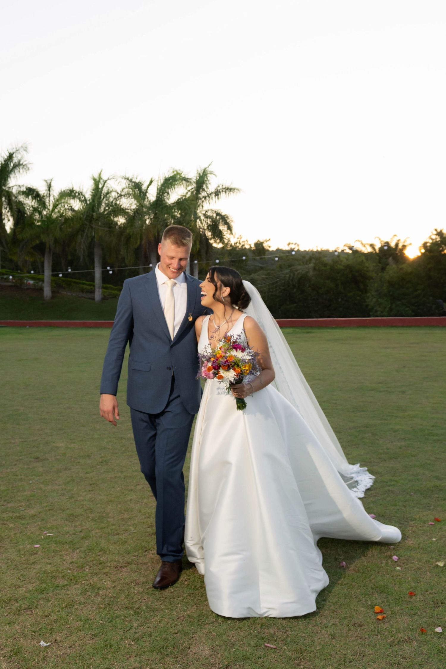 Bride and groom walking on green lawn with palm trees, she holds colorful bouquet in white gown, he wears blue suit.