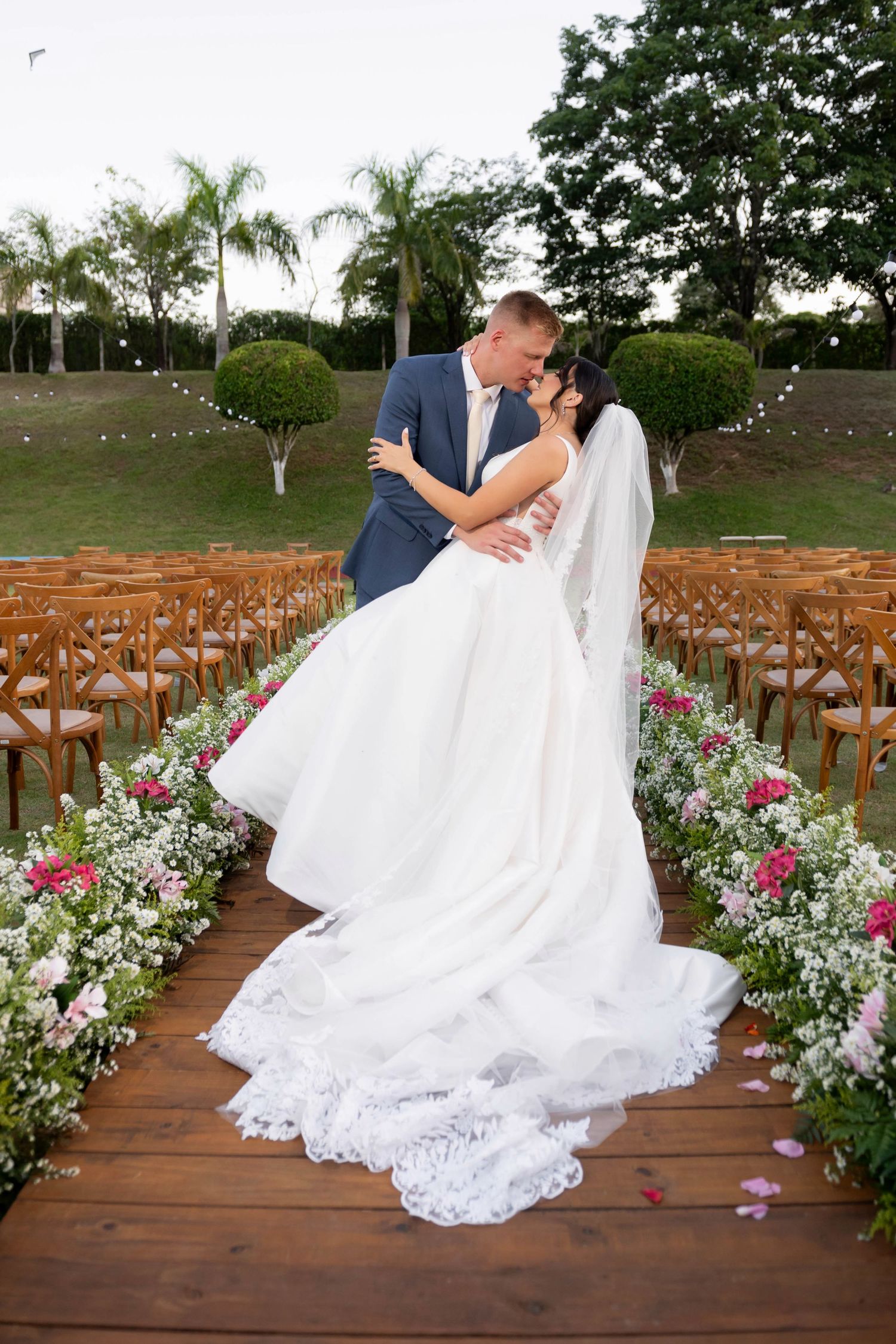 Bride and groom share a kiss at outdoor wedding ceremony aisle lined with pink and white flowers on wooden deck.