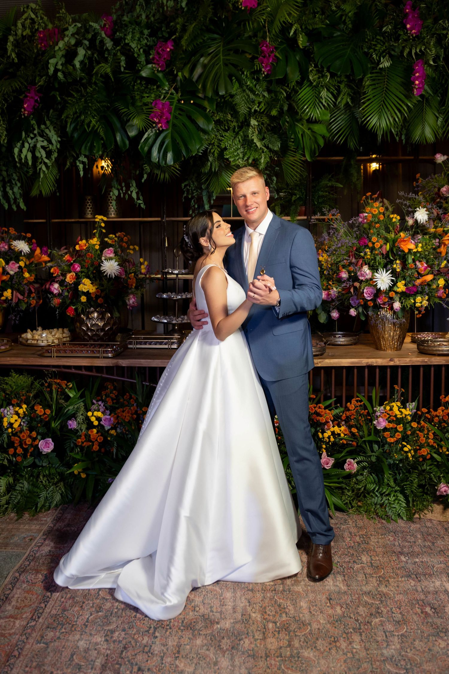 Bride in white gown and groom in blue suit pose together surrounded by lush tropical florals at wedding reception.