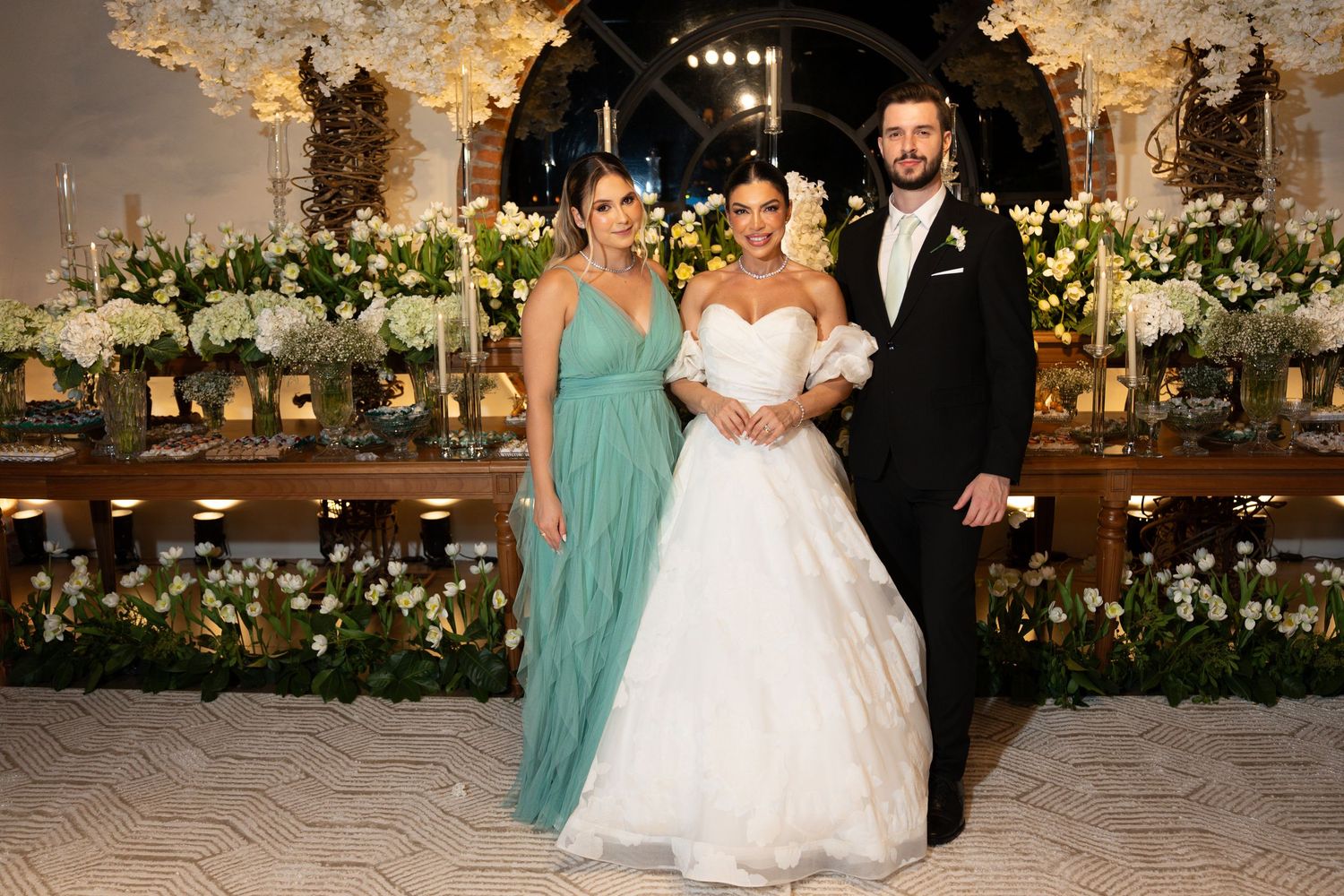 Bride in white gown poses with groom in tuxedo and bridesmaid in teal dress at elegant floral wedding reception.