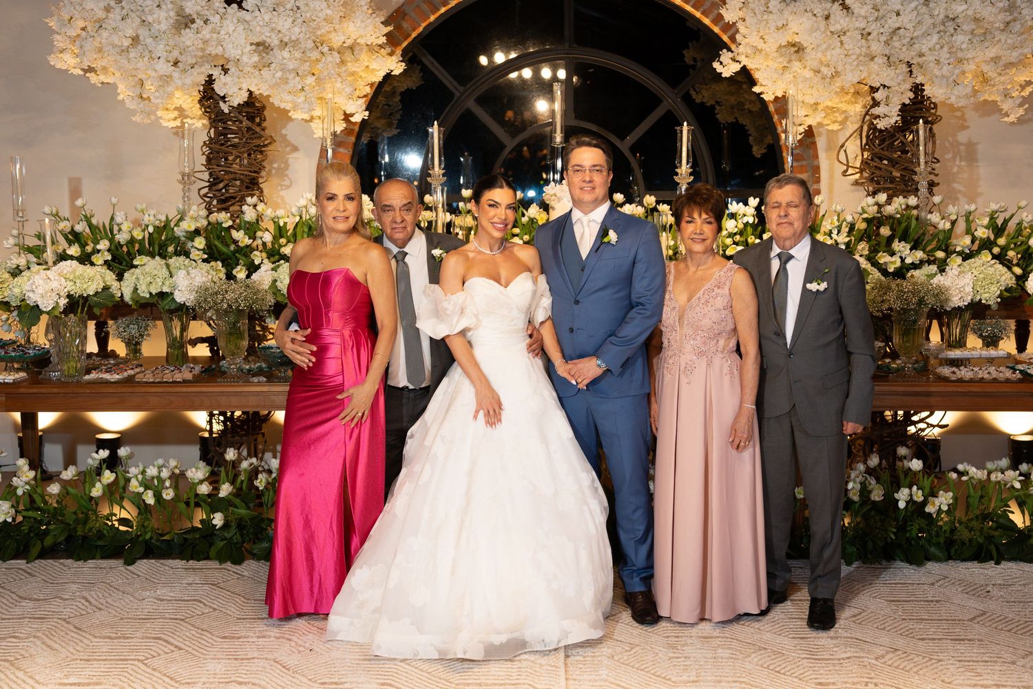 Wedding couple in white gown and blue suit pose with family members before floral backdrop at elegant reception.