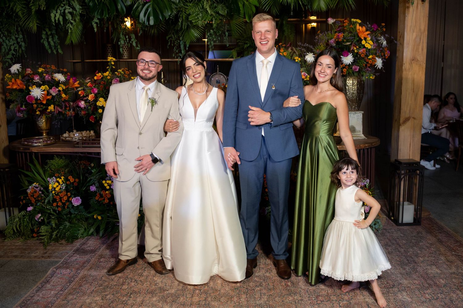 Wedding party posing together, bride in white gown, groom in blue suit, guests in green dress and tan suit, small flower girl.