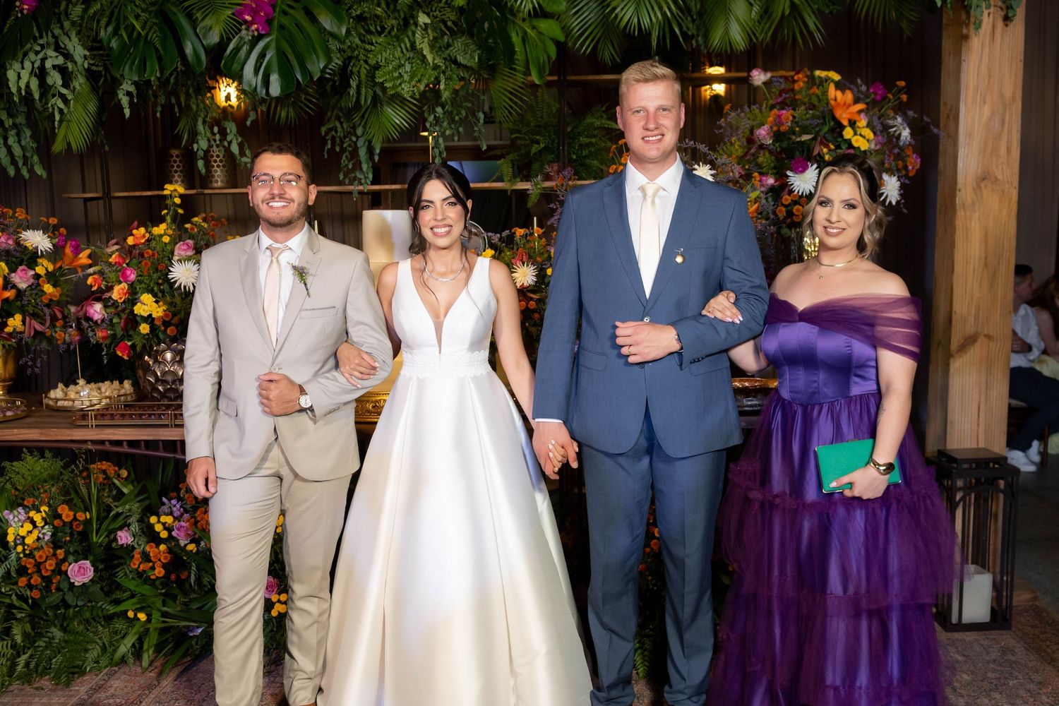 Wedding party of four posing indoors with lush greenery and colorful floral decor in the background.