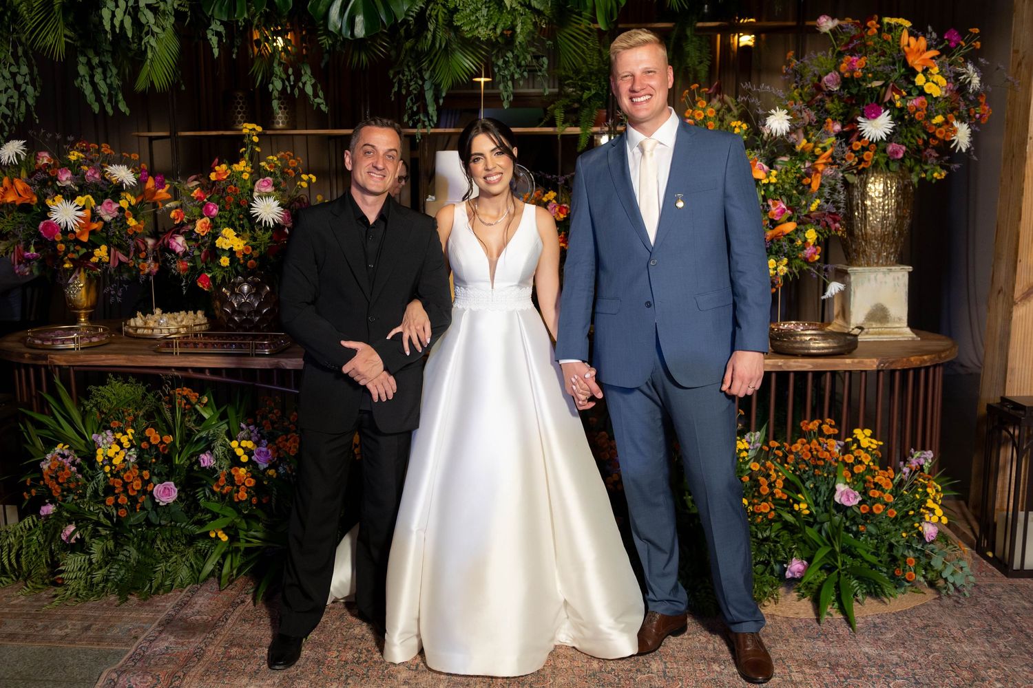 Bride in white gown stands between two men in formal attire, surrounded by lush floral arrangements at a wedding reception.