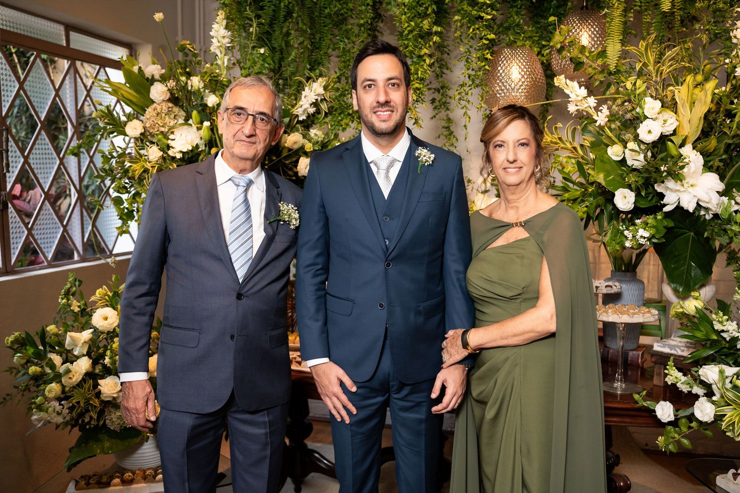 Three elegantly dressed people posing together at a floral wedding reception with lush green and white flower arrangements.