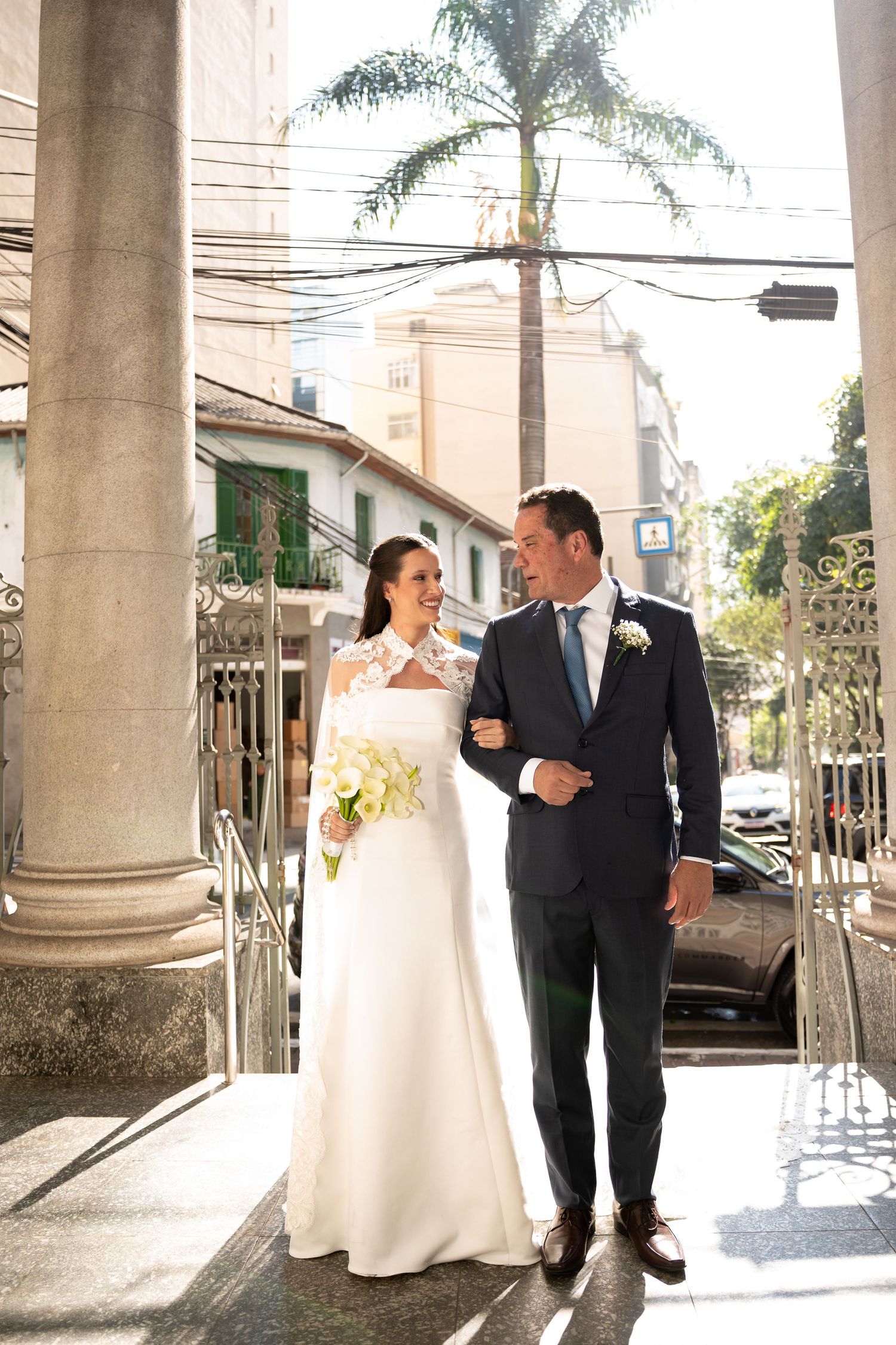 Bride and groom walk arm in arm outside a church, she holds white flowers, he wears a navy suit with boutonniere.