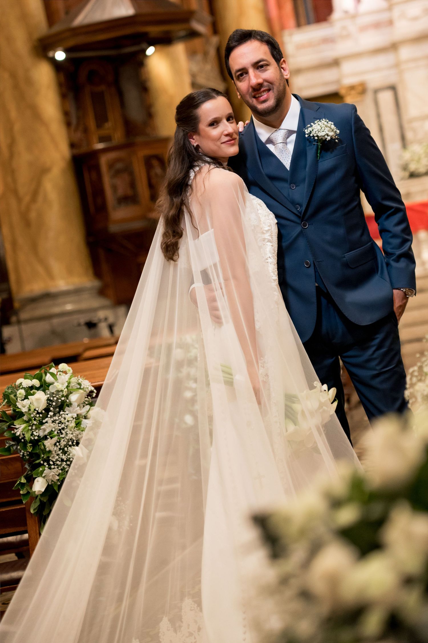 Bride in white gown with veil and groom in blue suit pose together inside an elegant church after wedding ceremony.
