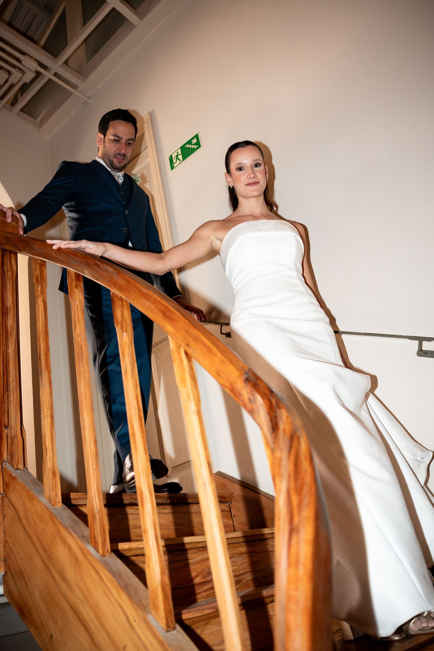 Bride in white gown and groom in navy suit pose elegantly on wooden staircase at wedding venue.