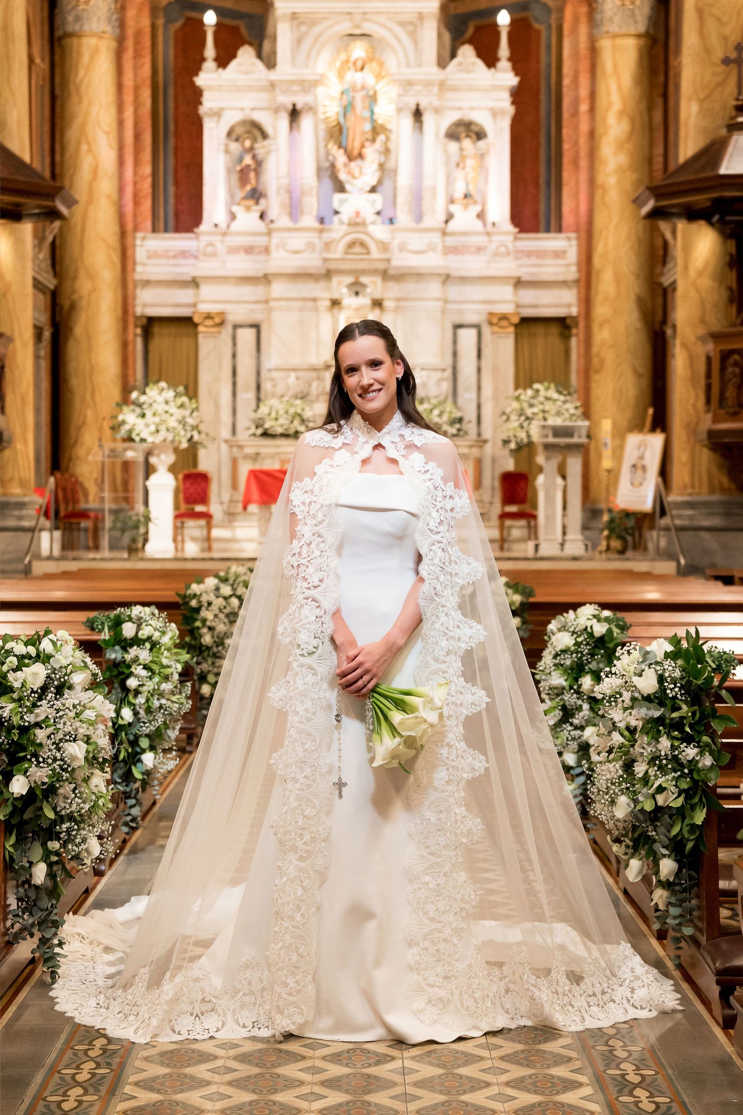 Bride in white gown and lace cape holds bouquet at church altar adorned with white floral arrangements.