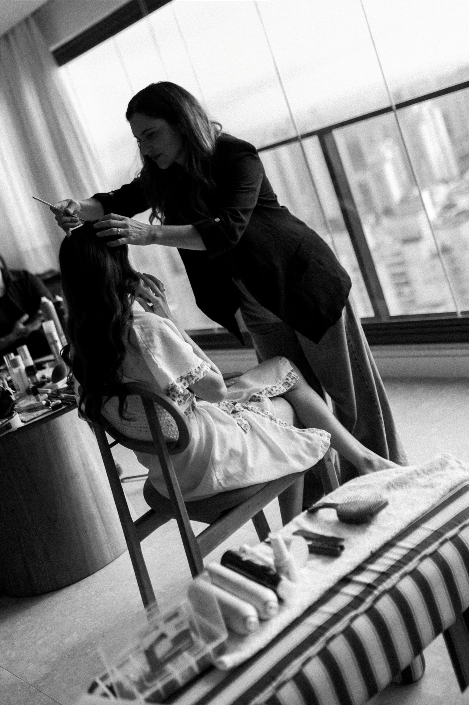 Black and white photo of a hairstylist working on a seated client near large windows in an elegant room.