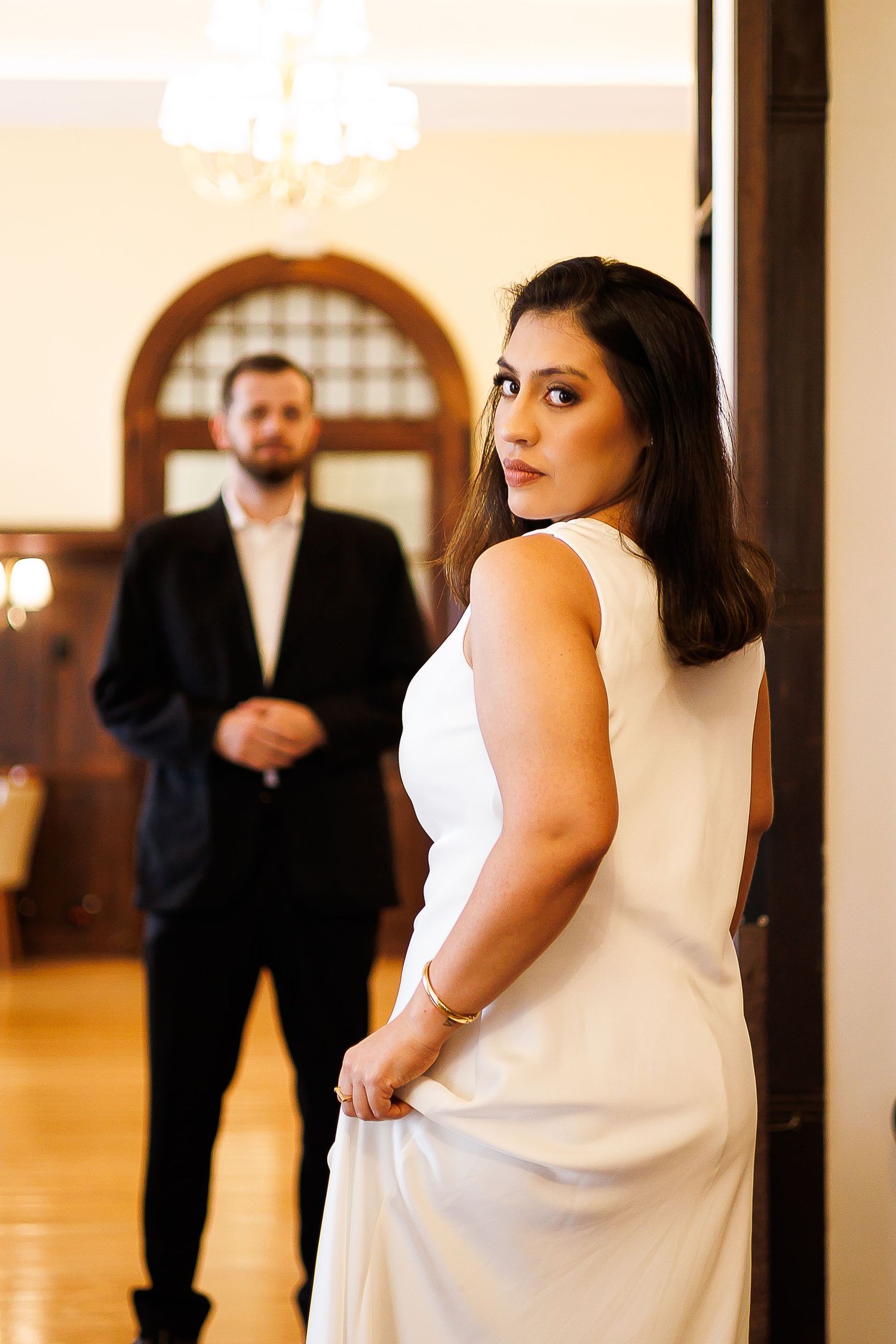 Bride in white gown looks back at groom in black suit in elegant venue with chandelier.