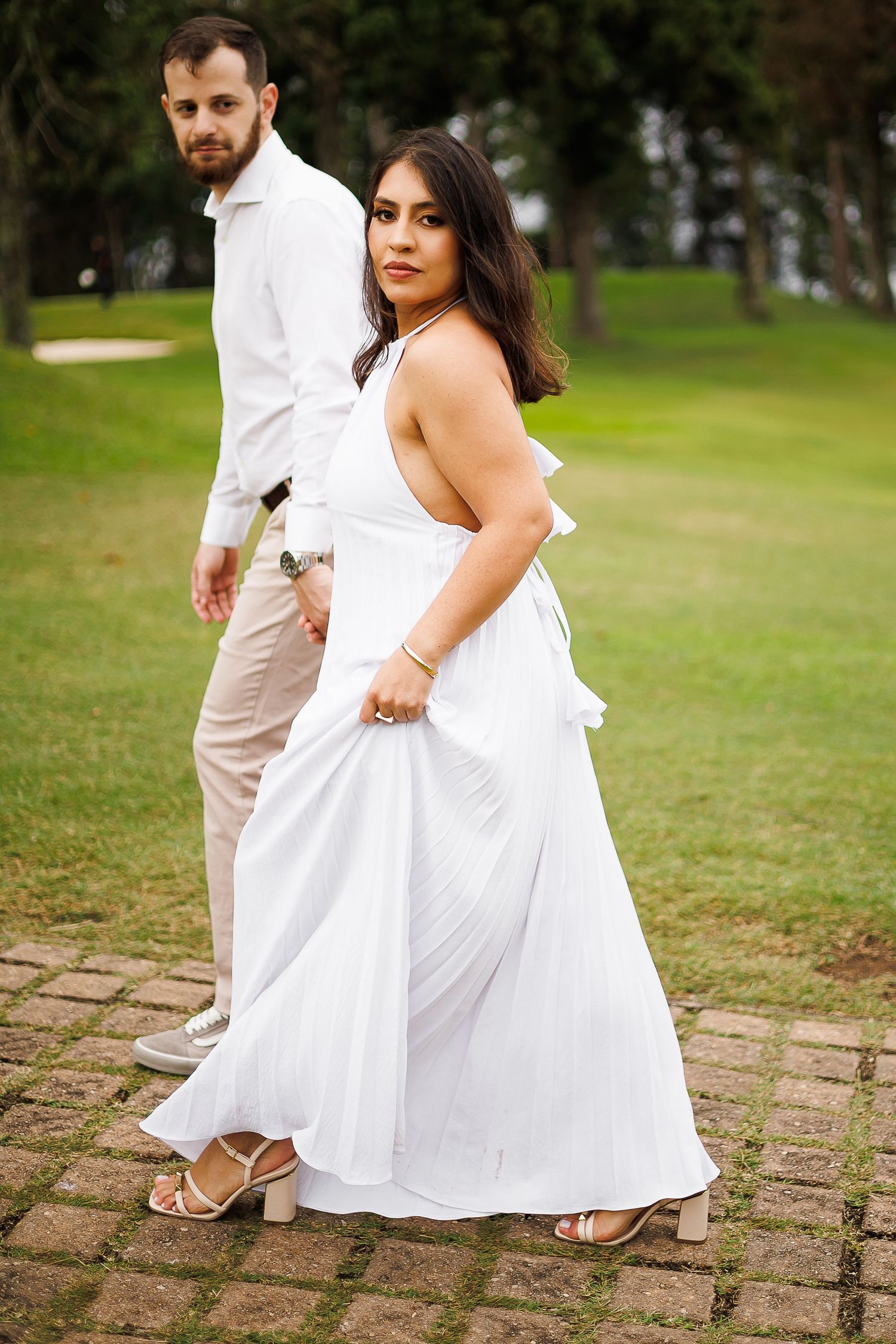 Couple in white outfits posing on stone path in lush green park, woman in elegant white gown looks back gracefully.