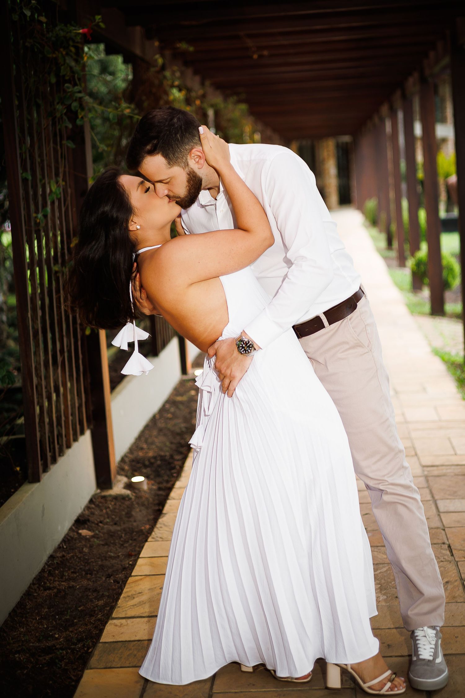 Couple in white outfits sharing a romantic kiss while embracing on a sunlit garden pathway.