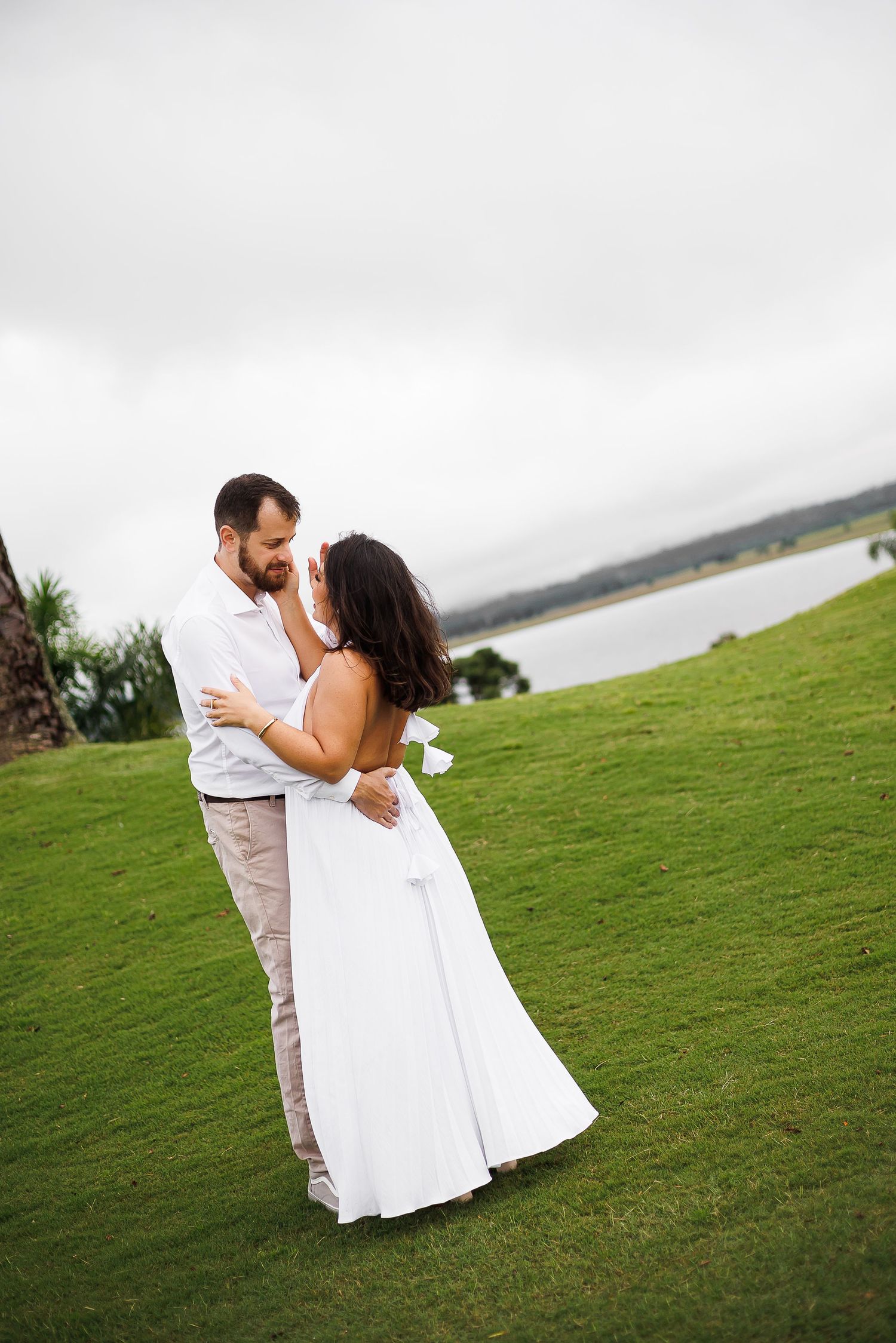 Bride and groom embrace on green lawn near lake, wearing white wedding attire on overcast day.