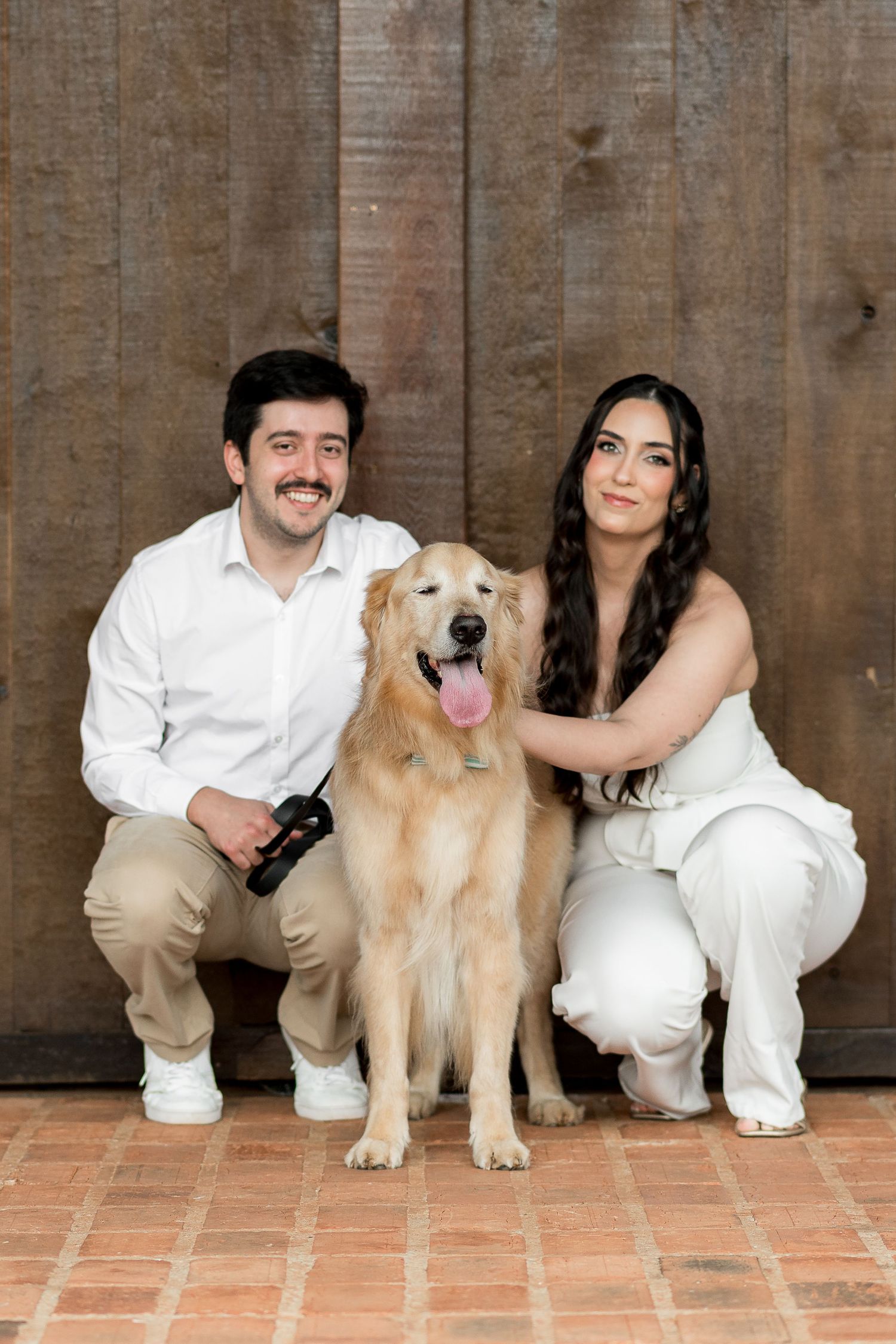 Couple in white outfits crouching beside a happy Golden Retriever dog in front of a rustic wooden wall.