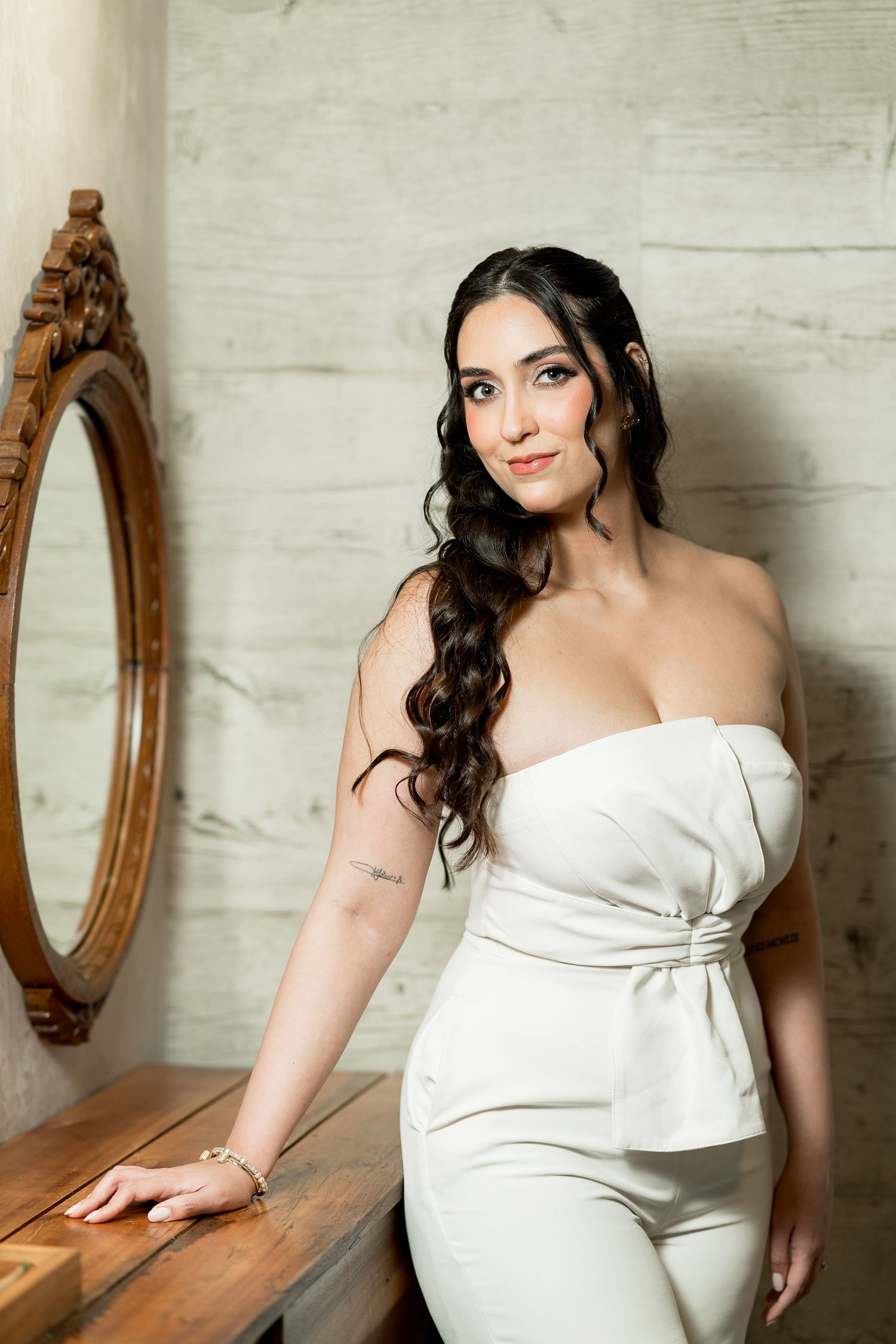 Elegant woman in white strapless jumpsuit posing by vintage mirror and wooden dresser with long wavy dark hair.
