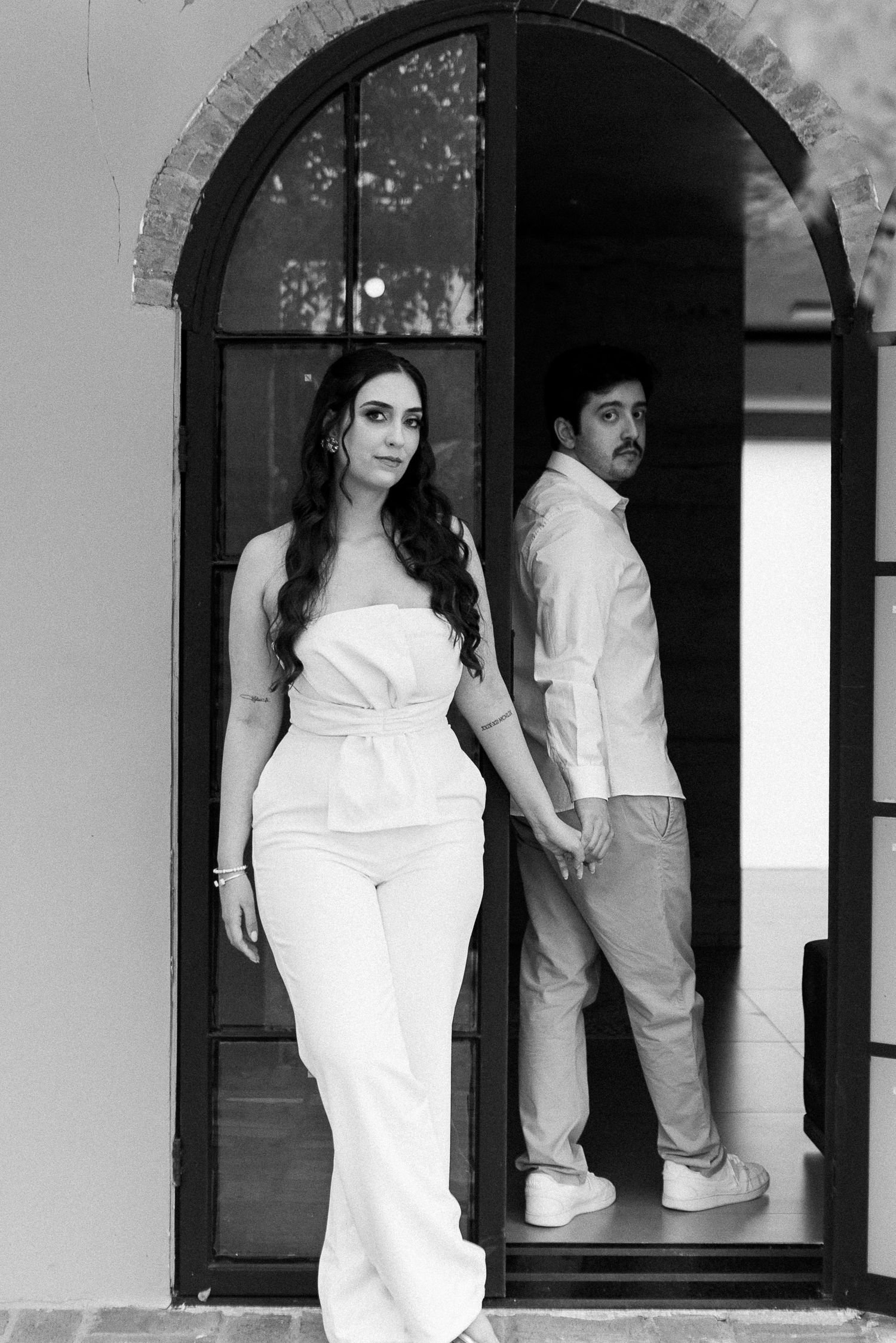 Black and white photo of an elegant couple posing in an arched doorway, woman in white jumpsuit, man in casual attire.