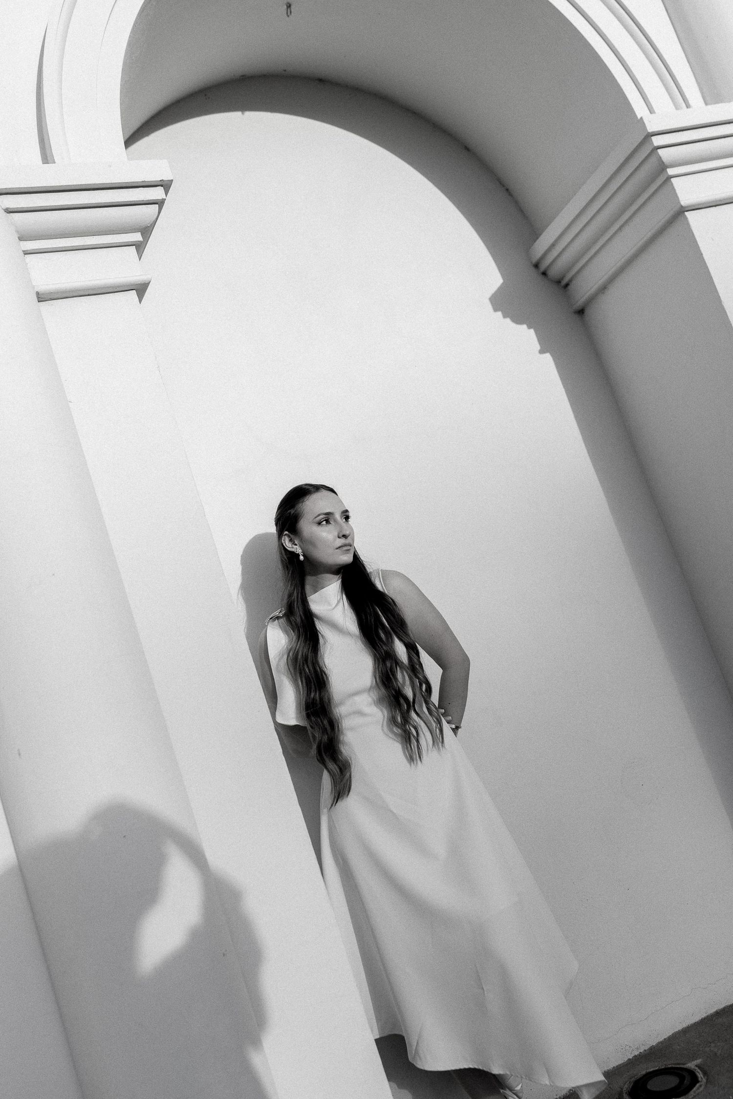 Black and white photo of a young woman in a white dress posing under an elegant arch.