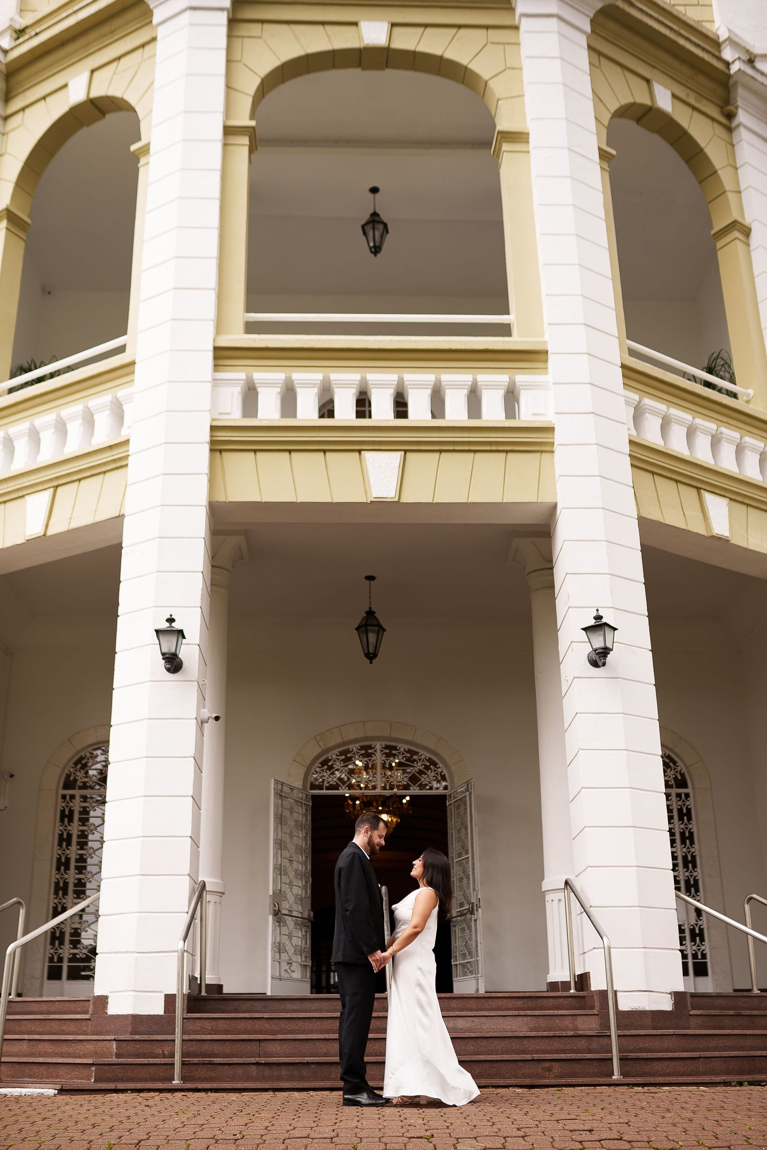 Bride and groom standing together in front of elegant white colonial building with arched windows and tall columns.