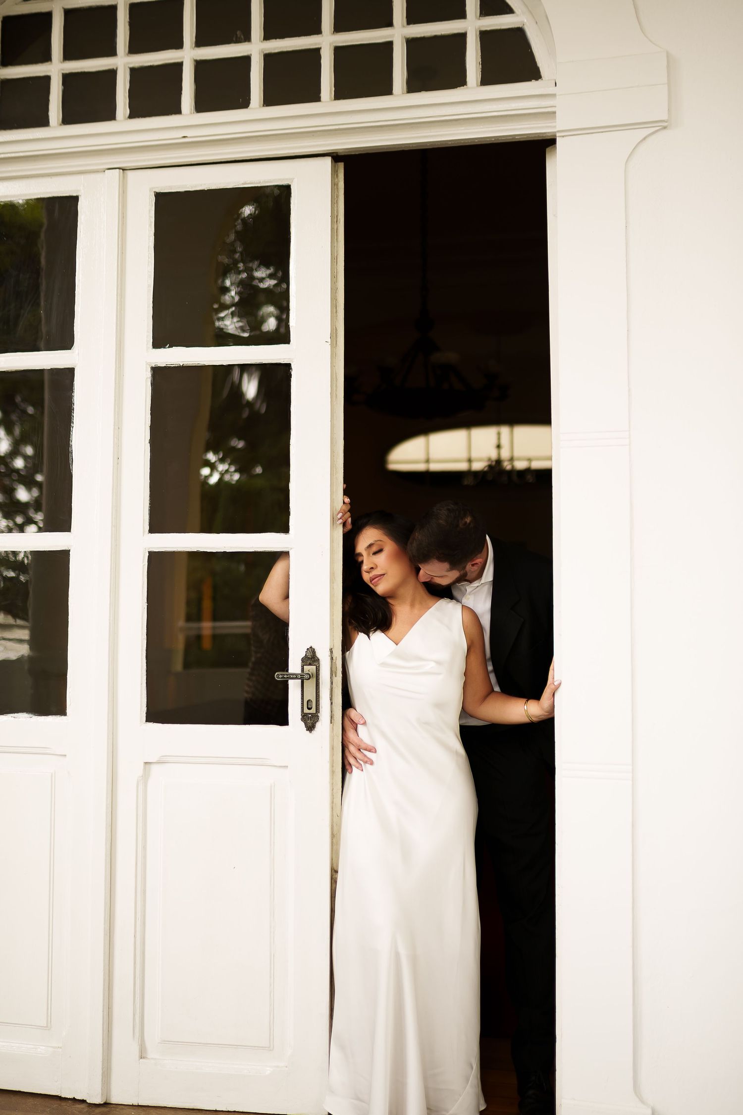 Bride in elegant white satin gown stands in French doors as groom embraces her from behind.