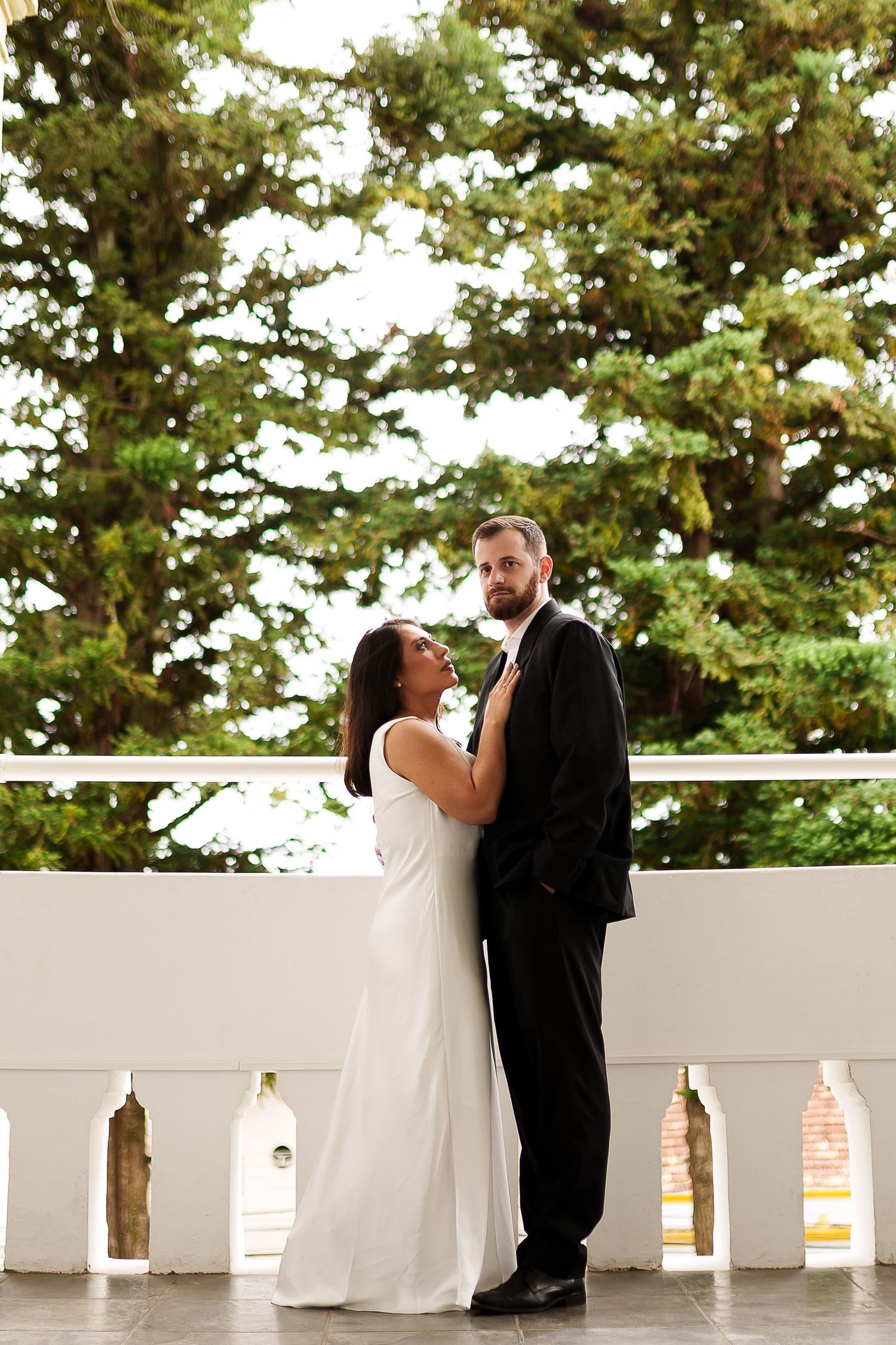 Bride in white gown and groom in black suit embrace on a balcony with lush green trees in the background.