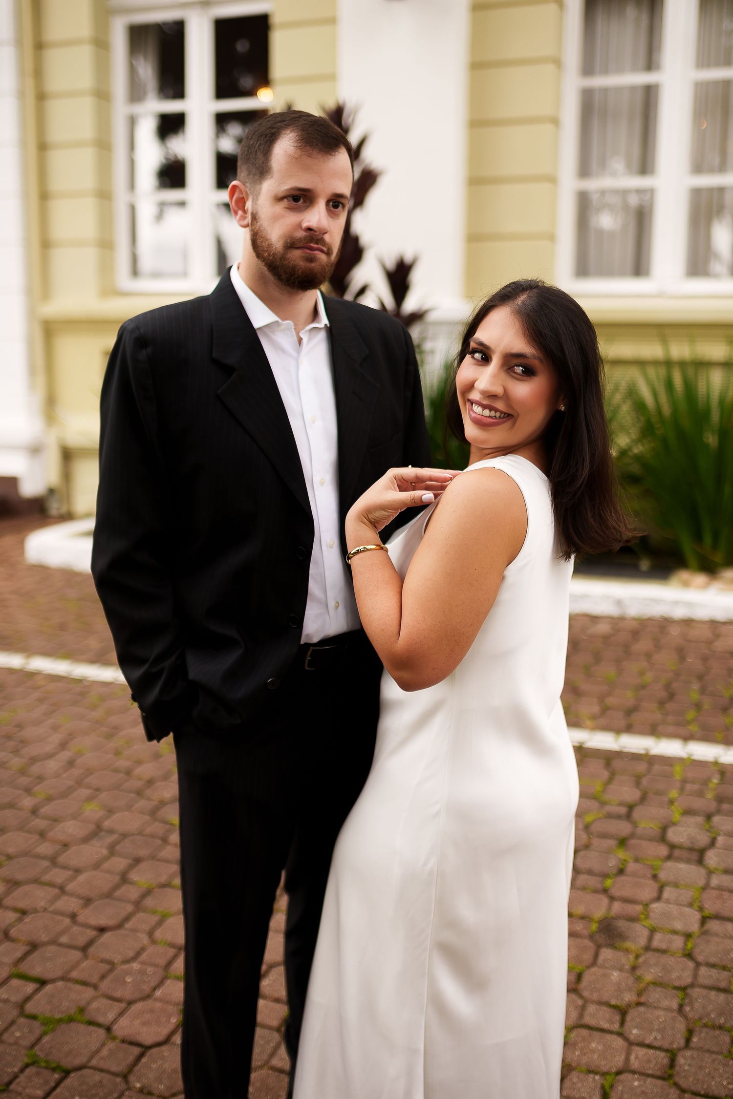 A smiling couple in formal attire, woman in white dress and man in black suit, posing outdoors near a building.
