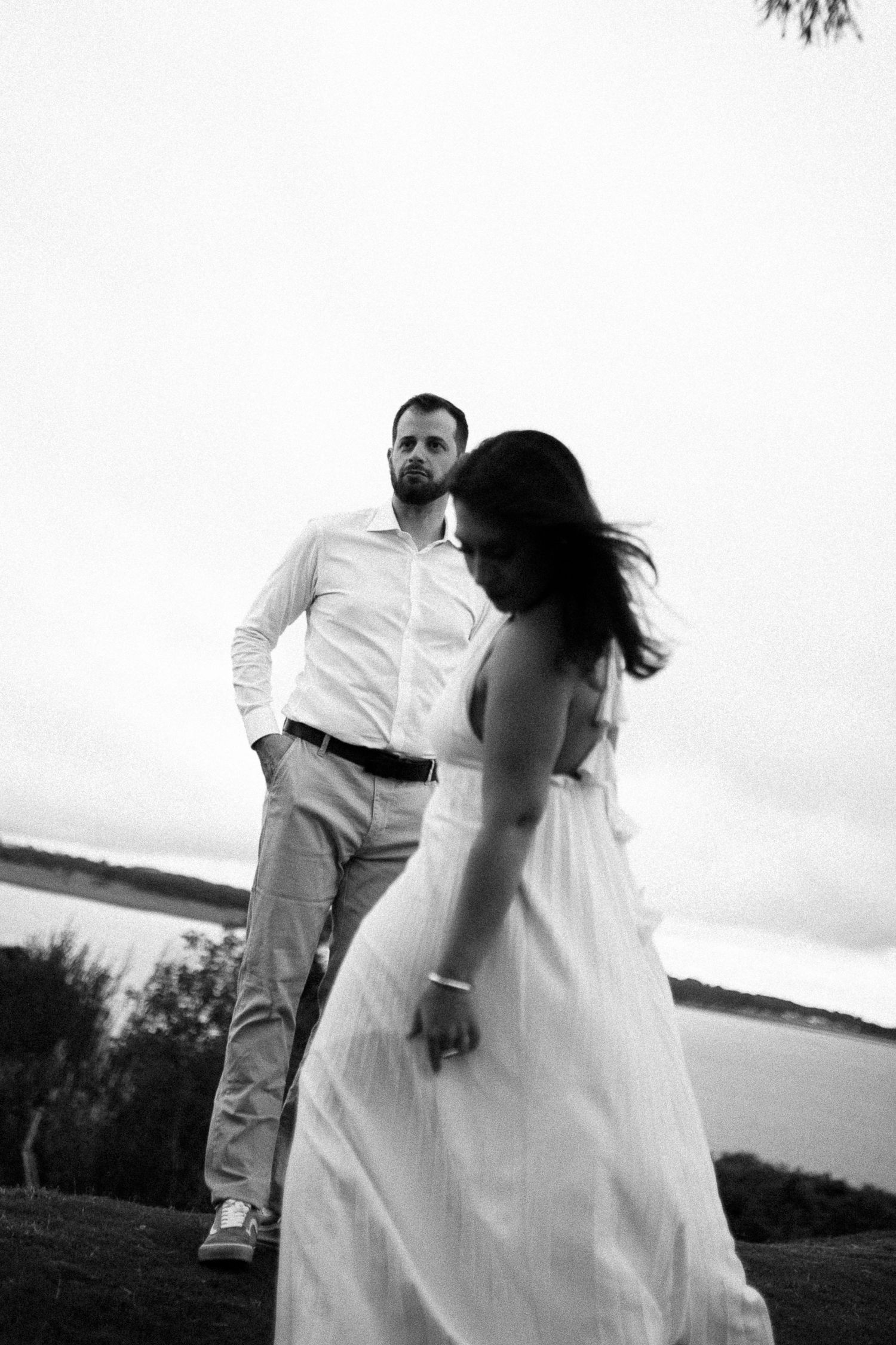 Black and white photo of a couple outdoors near water, woman in white dress with wind-blown hair, man in white shirt.