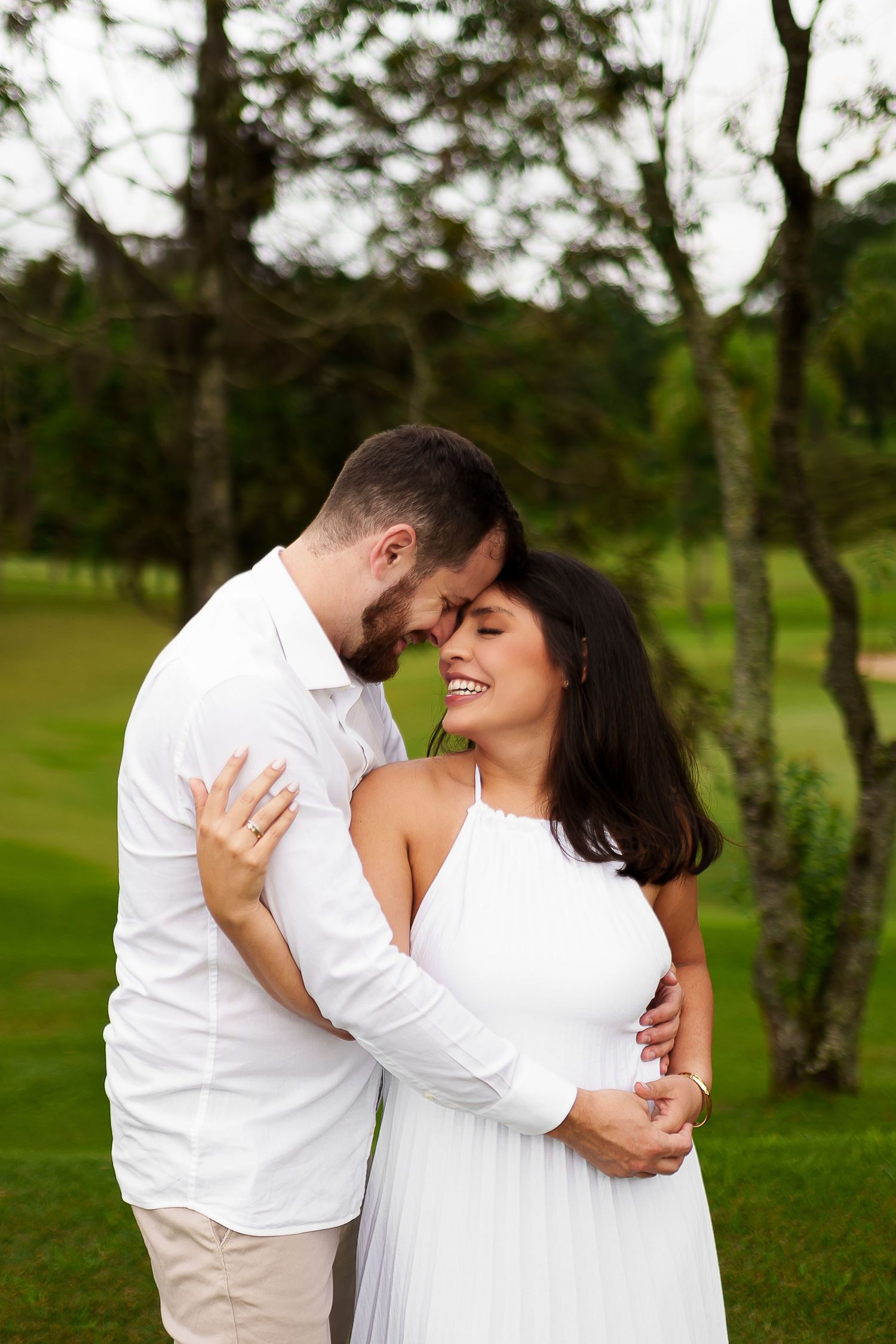 A couple in white outfits embrace and smile in a lush green outdoor setting, sharing a romantic moment.