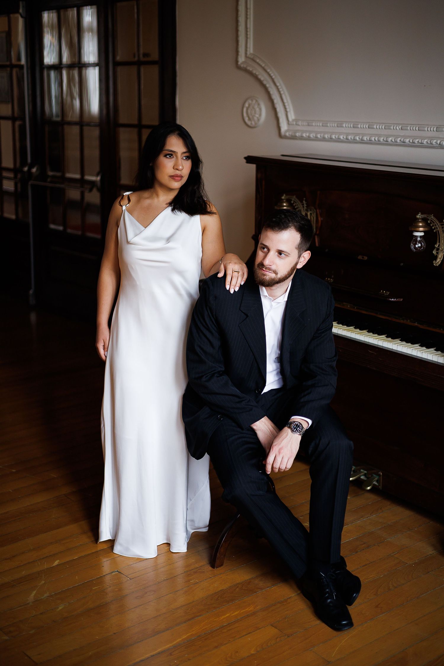 Elegant couple posing by a piano, woman in white gown and man in black suit, in a classic interior setting.