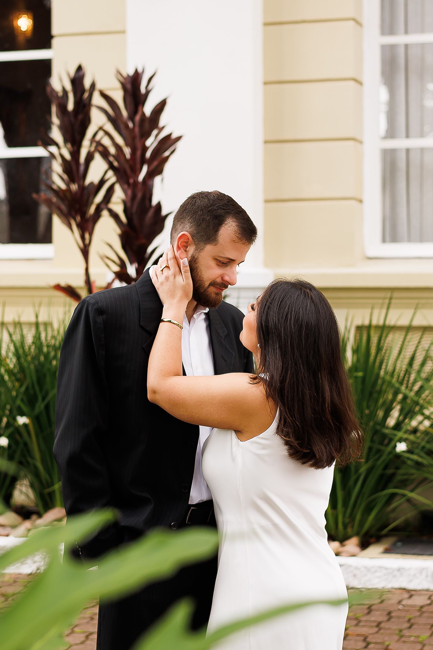 A couple in wedding attire share a romantic embrace outdoors, with the bride in white touching the groom's face tenderly.