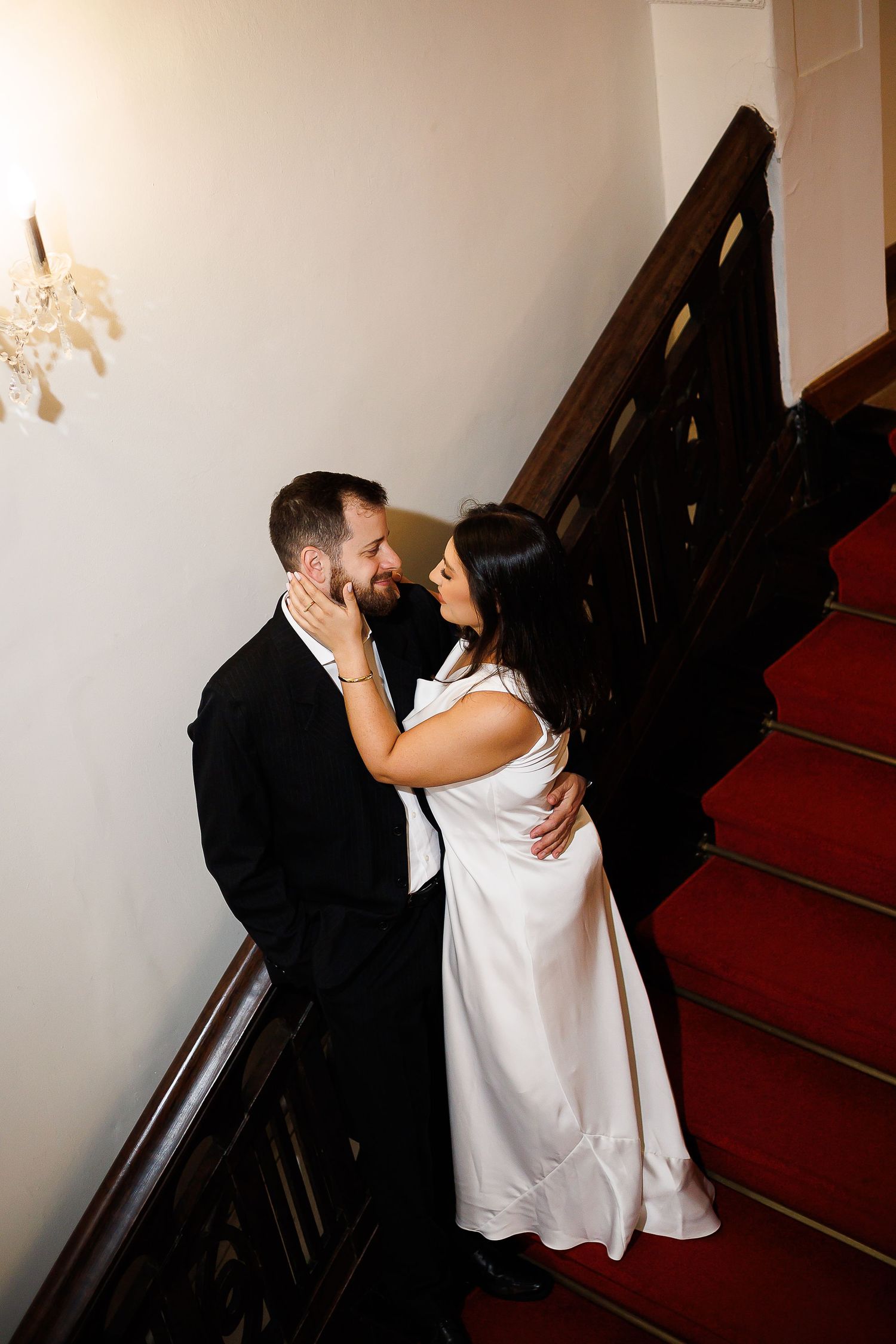 Bride and groom share a romantic moment on a grand staircase with red carpet, dressed in wedding attire.