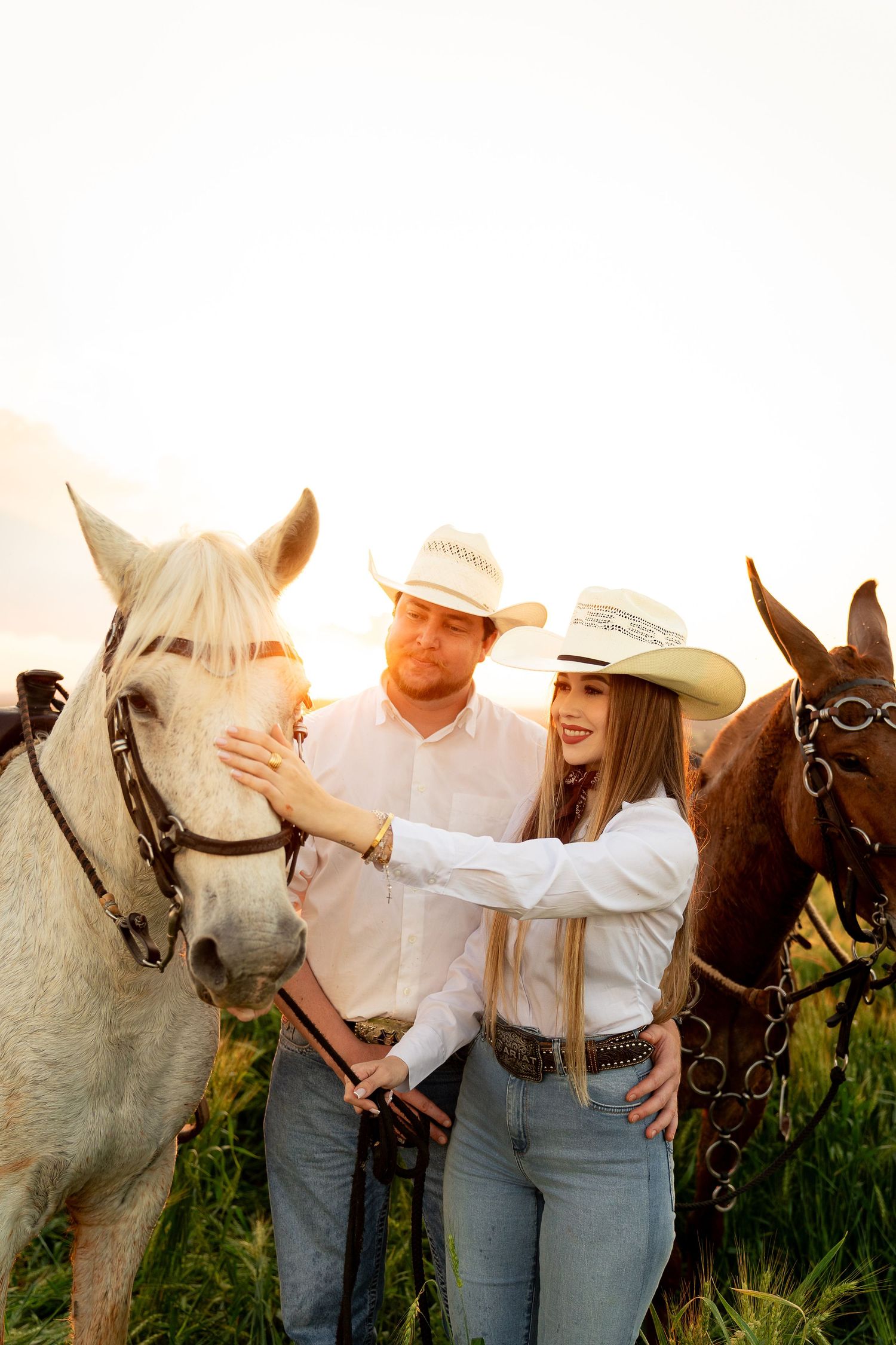 A couple in cowboy hats and white shirts stand with two horses at sunset in a grassy field.