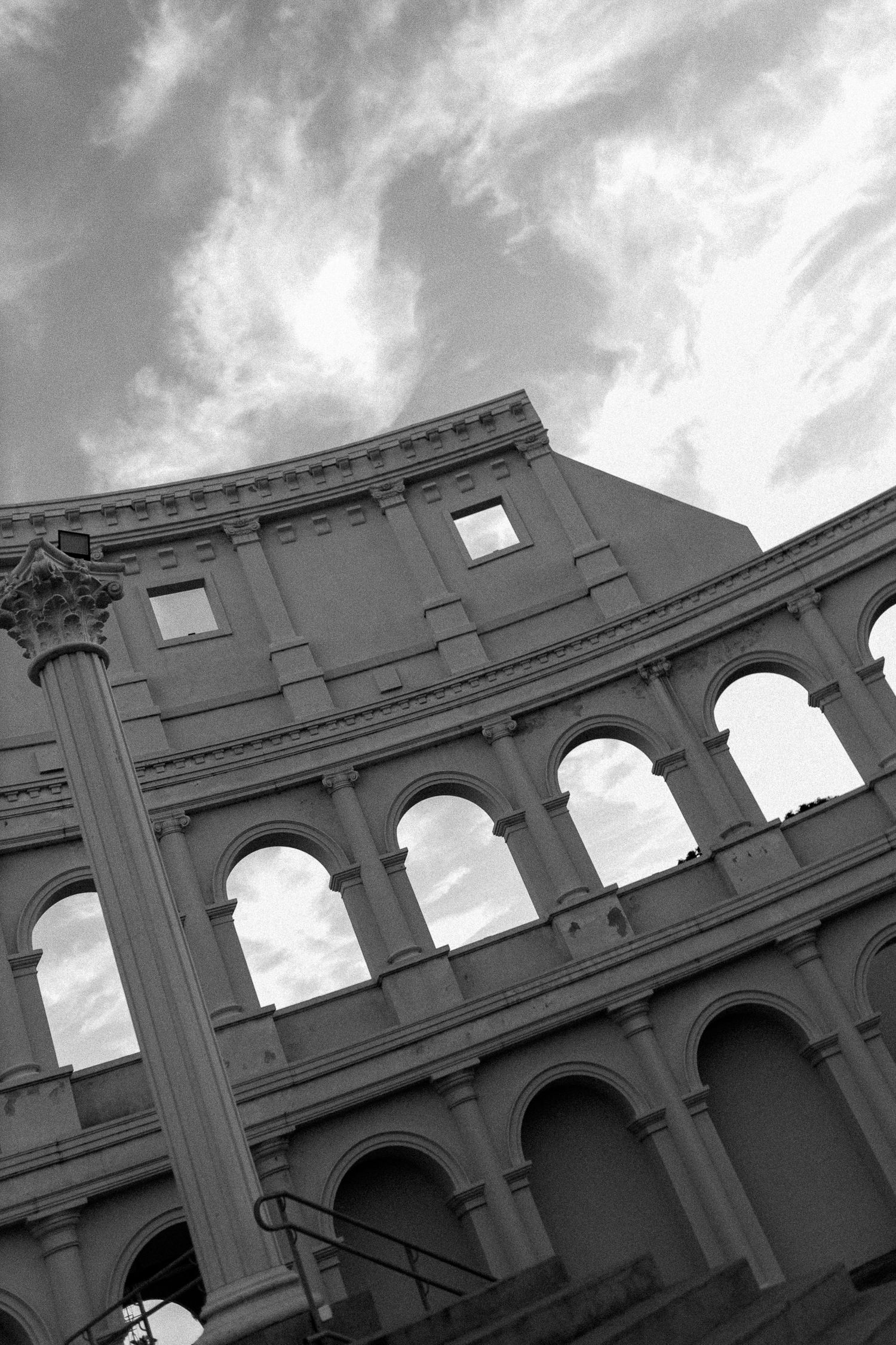 Black and white photo of the Roman Colosseum facade with arched windows against a cloudy sky.