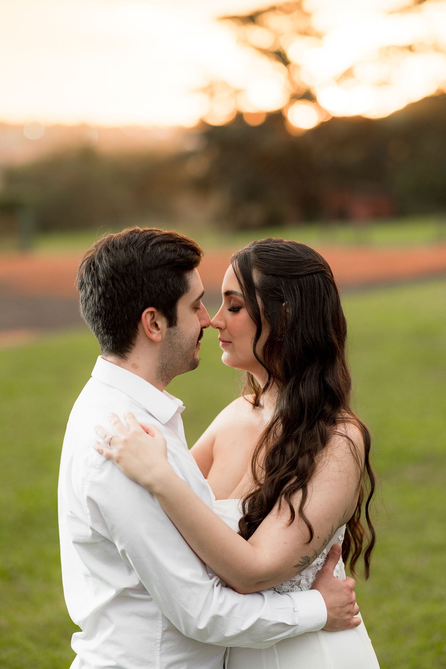 A couple in white attire shares a romantic embrace at golden hour with a lush green field backdrop.
