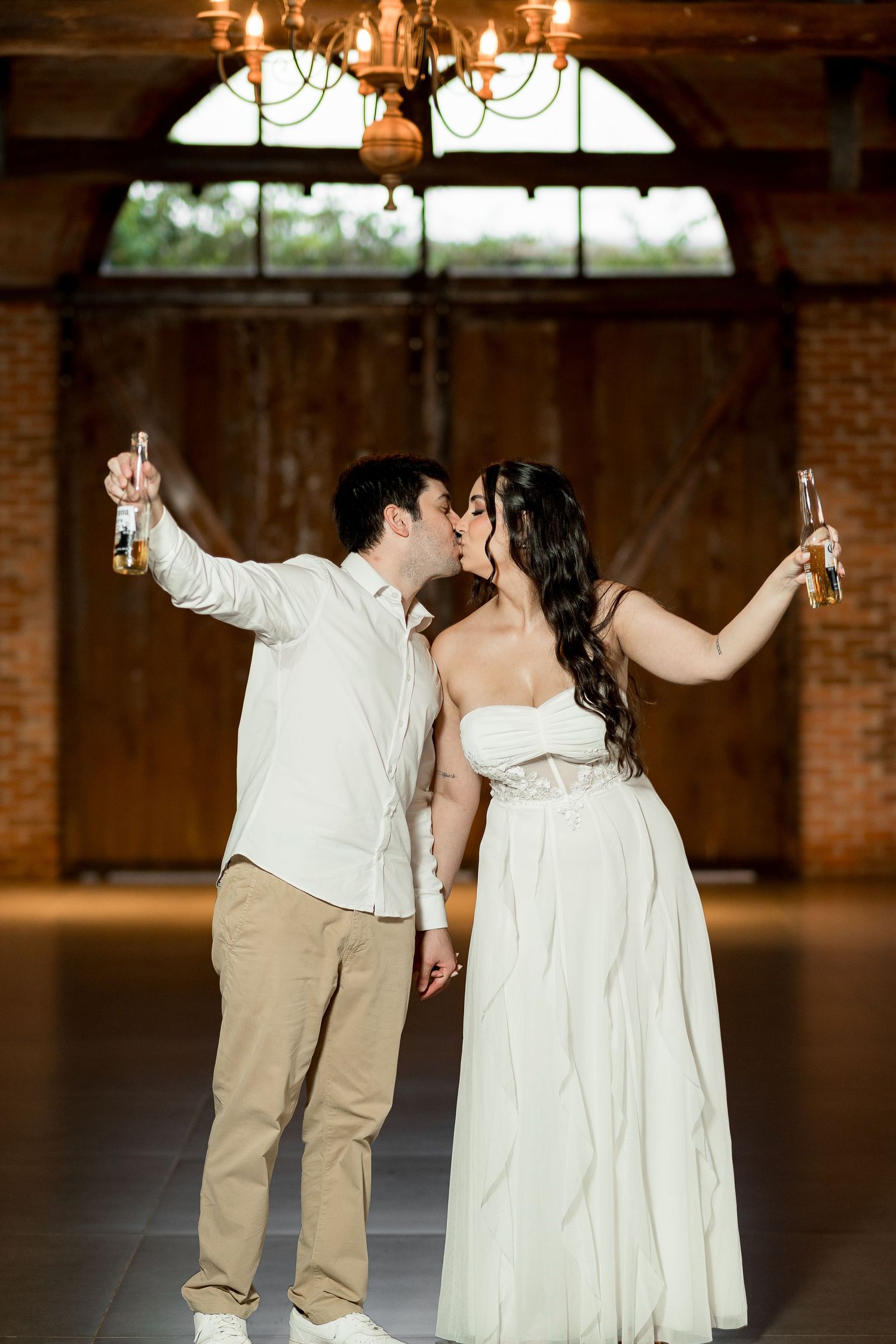 A couple in white attire kisses while raising beer bottles in a rustic barn venue with brick walls and chandelier.