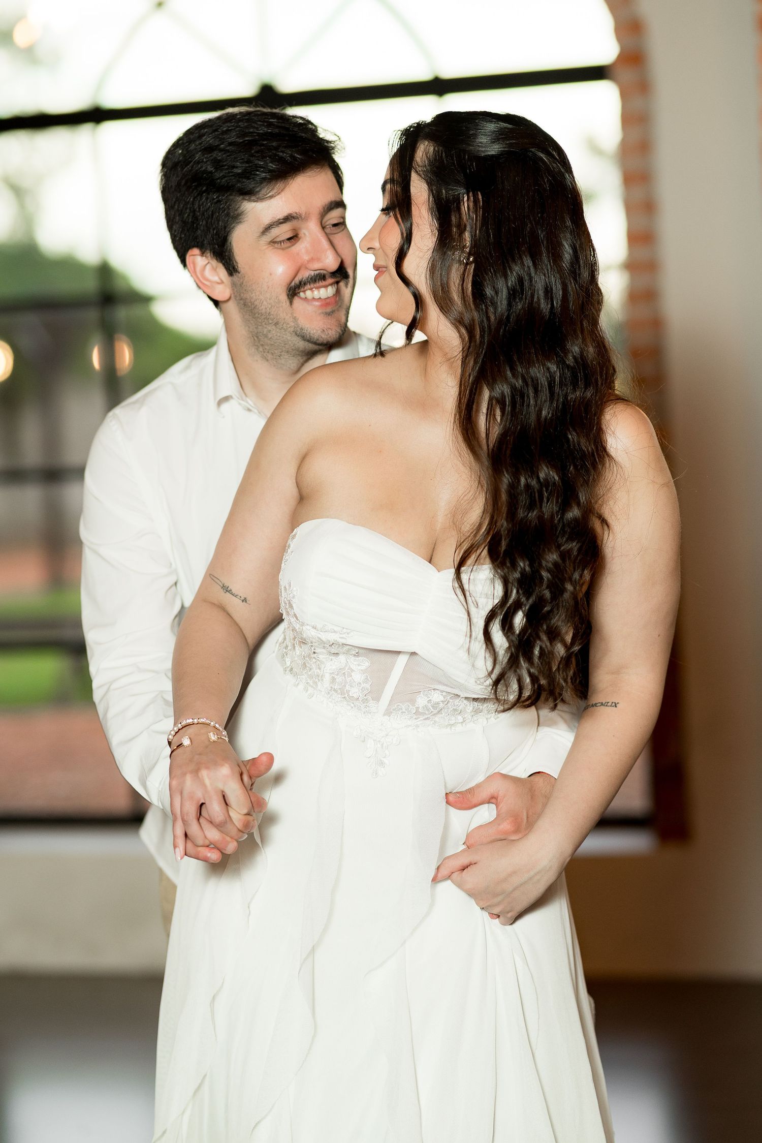 Smiling groom in white shirt embraces bride in strapless white gown during romantic first dance at wedding reception.