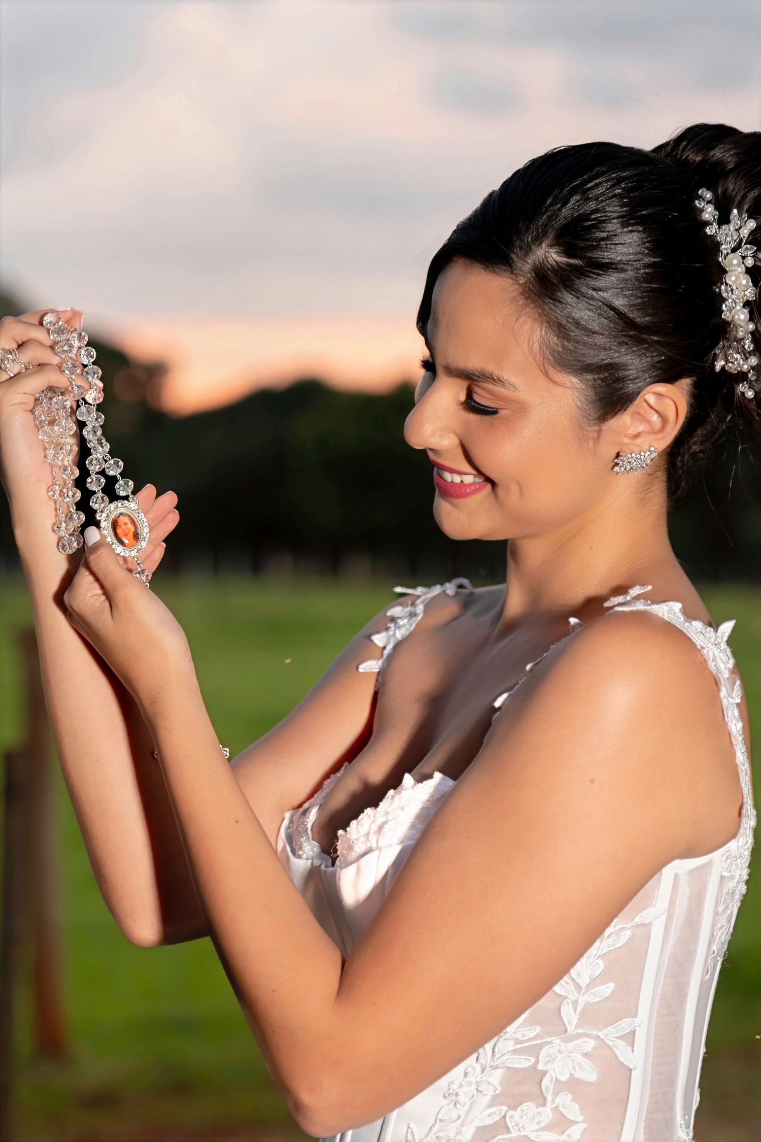 Smiling bride in white lace dress holds a rosary necklace outdoors at sunset with green fields behind her.