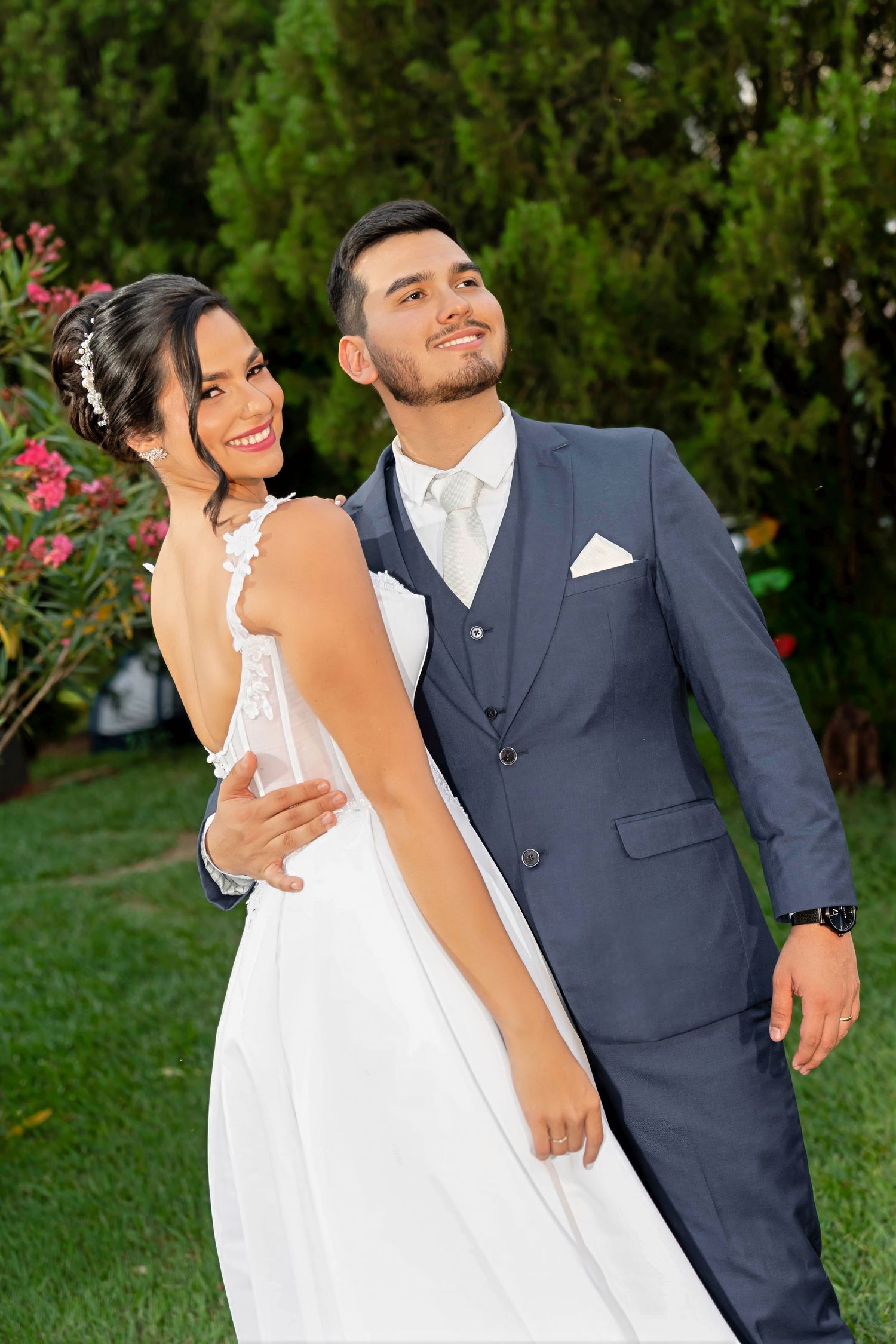A smiling bride in a white gown and groom in a blue suit pose together outdoors in a lush garden setting.