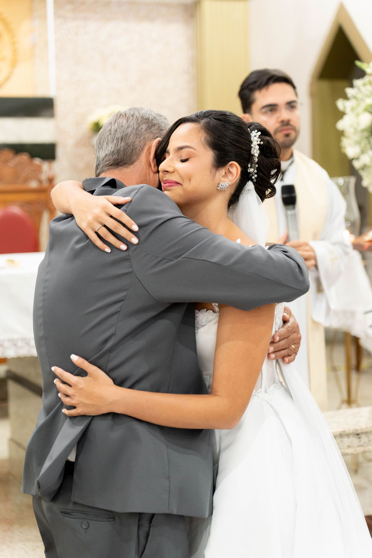 Bride in white gown shares emotional hug with man in gray suit during wedding ceremony in church.