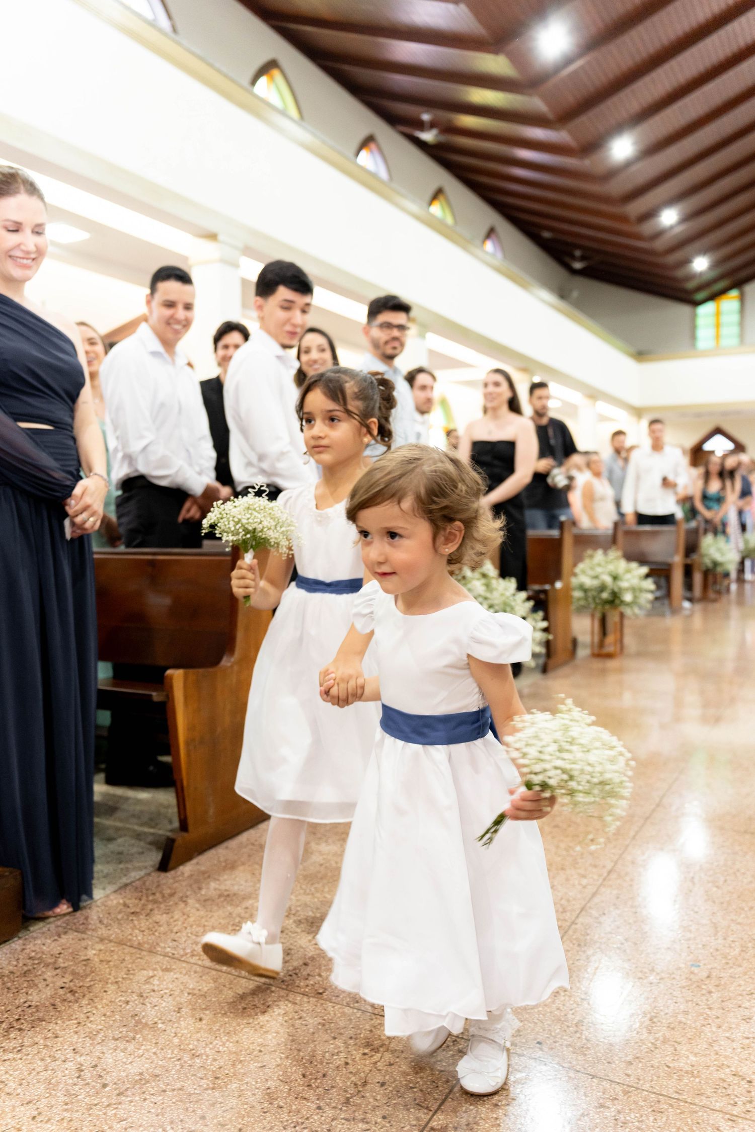 Two flower girls in white dresses with blue sashes walk down a church aisle holding bouquets at a wedding ceremony.