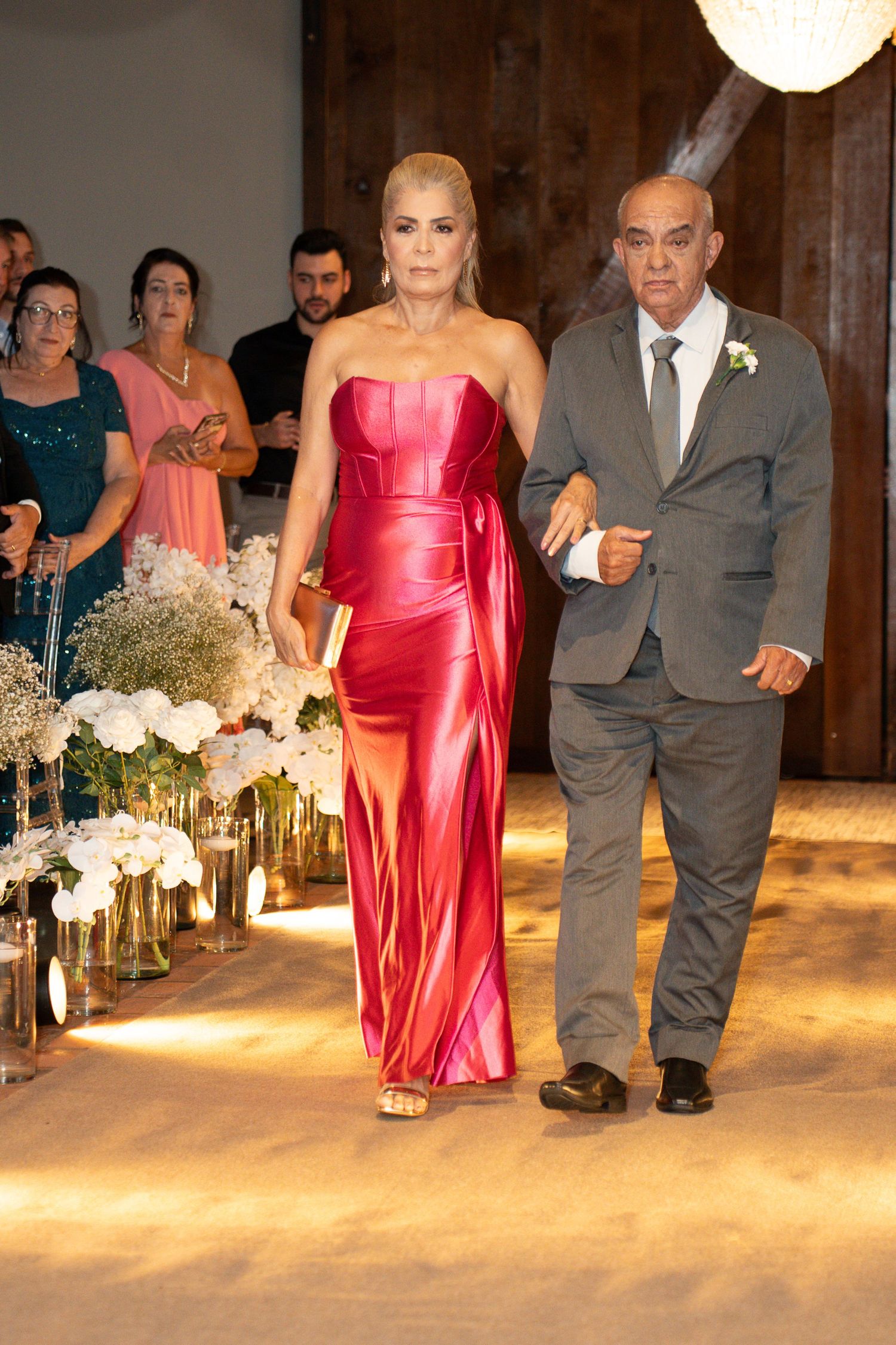 Woman in red satin gown walks down wedding aisle with older man in gray suit, flanked by floral arrangements.