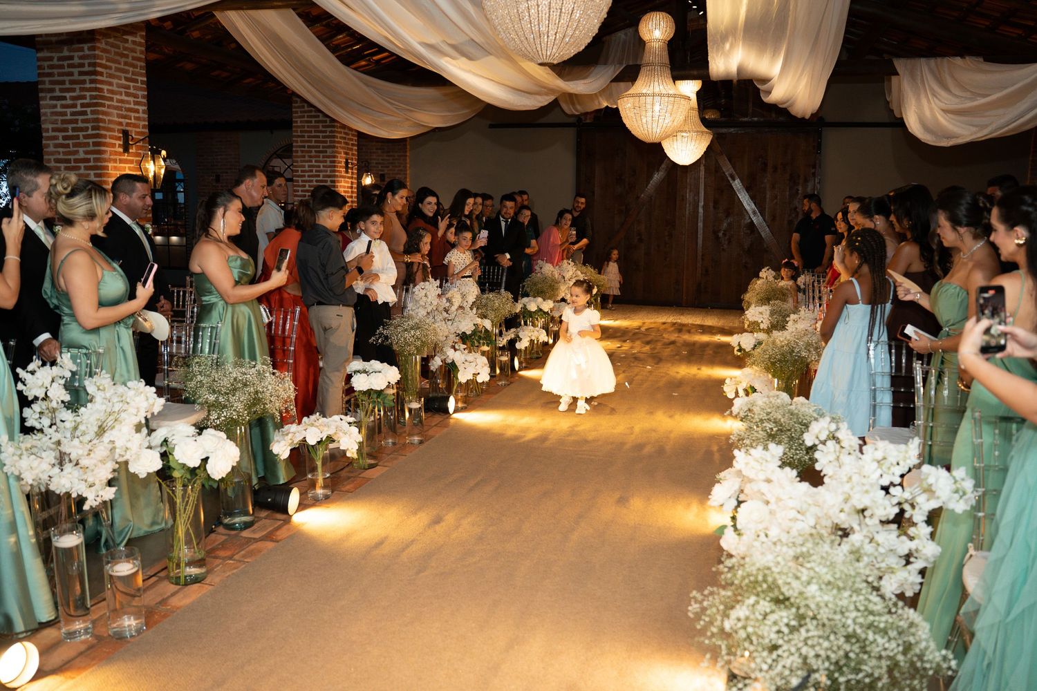A flower girl walks down a decorated wedding aisle flanked by bridesmaids in sage green dresses and white floral arrangements.